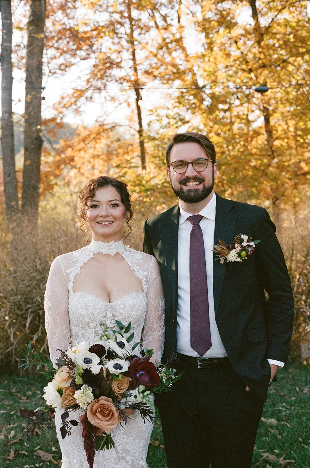 A bride and groom standing outdoors during fall, smiling at the camera. The bride wears a lace wedding dress, holding a large bouquet of flowers. The groom wears a black suit with a white shirt, purple tie, and boutonniere. Trees with orange and yellow leaves are in the background.