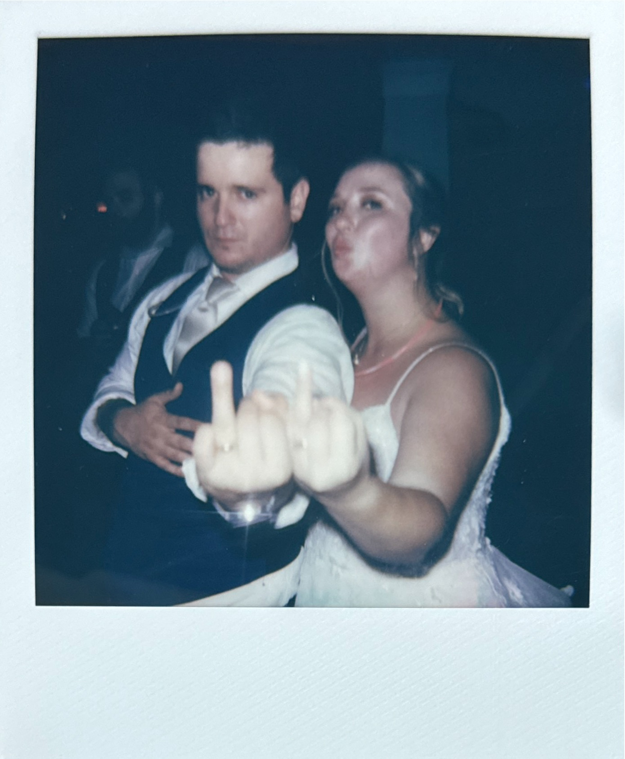 A couple dressed in formal attire posing playfully, with the woman showing her middle finger to the camera.