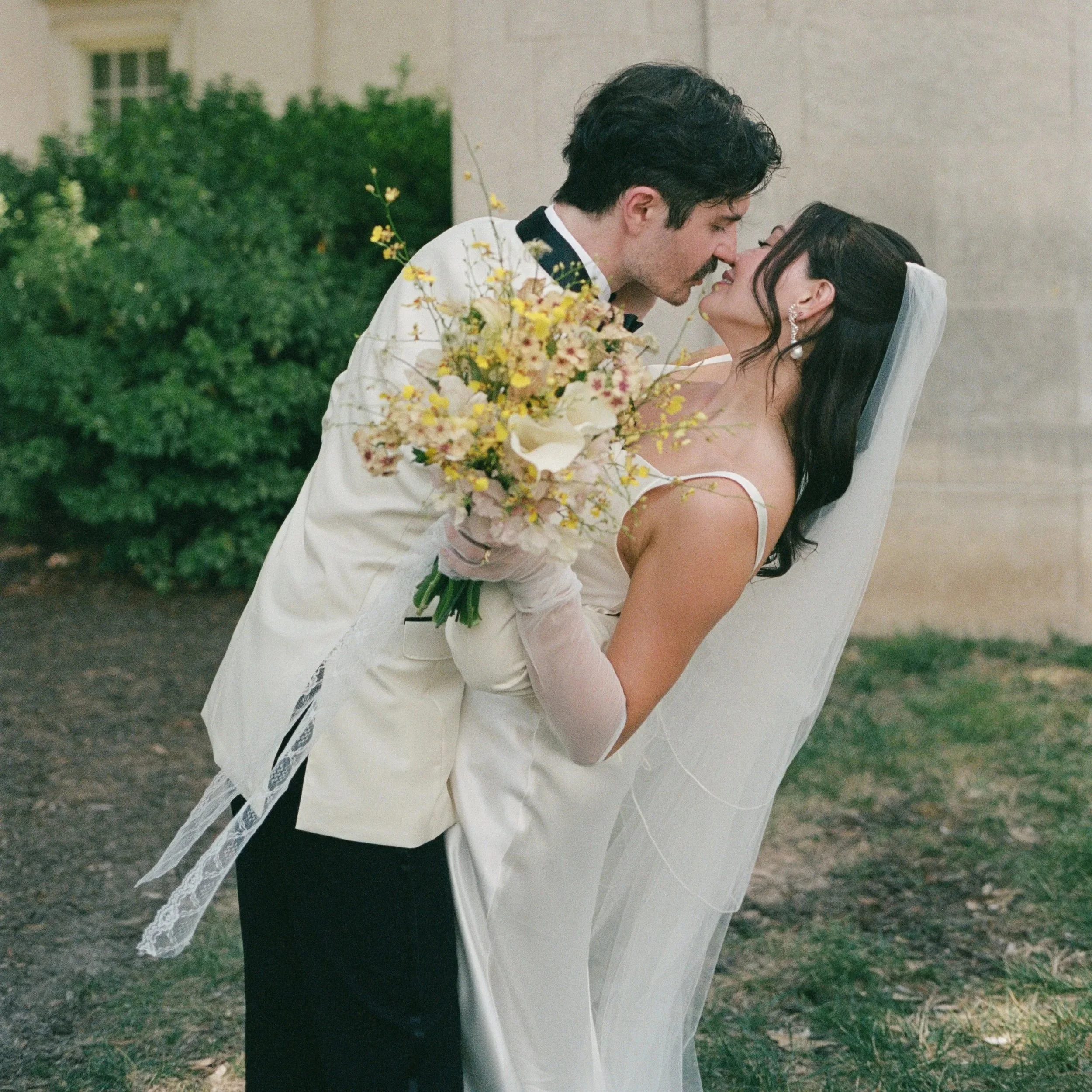 A groom and bride share a kiss outdoors during their wedding, with the groom wearing a white tuxedo and the bride in a white wedding dress holding a bouquet of flowers. Richmond Virginia Wedding Common House Film Photographer