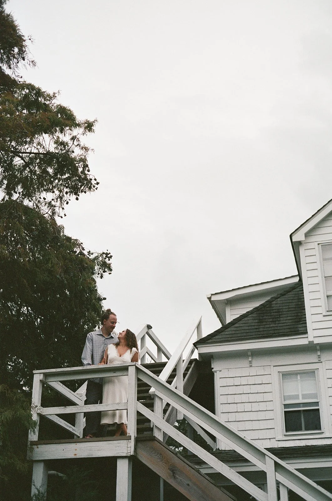A couple standing on a white wooden deck outside a white house, looking into each other's eyes under an overcast sky. ocracoke outerbanks wedding film photographer