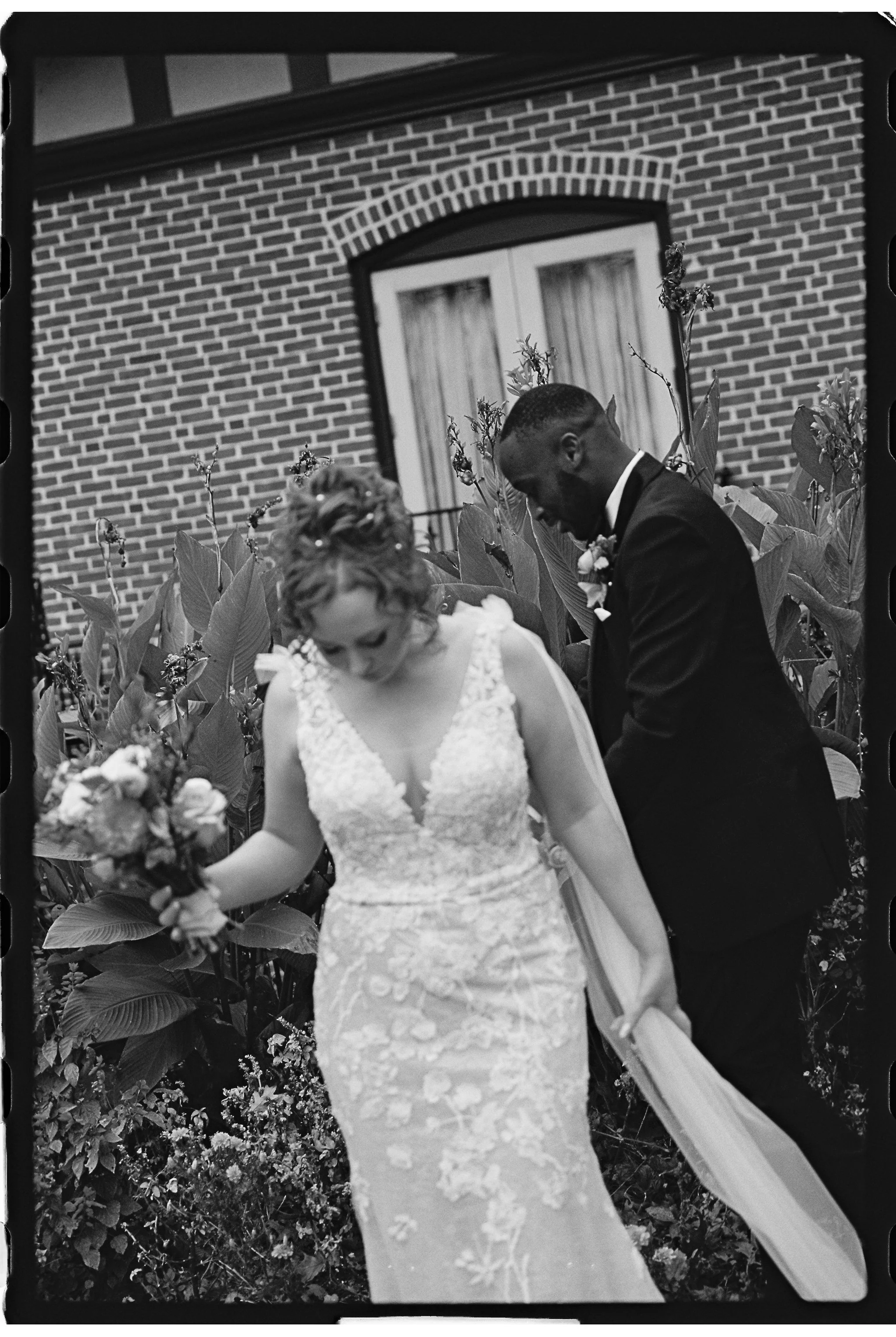 Bride in lace gown holding bouquet beside groom in suit by brick wall and plants. gramercy mansion maryland wedding