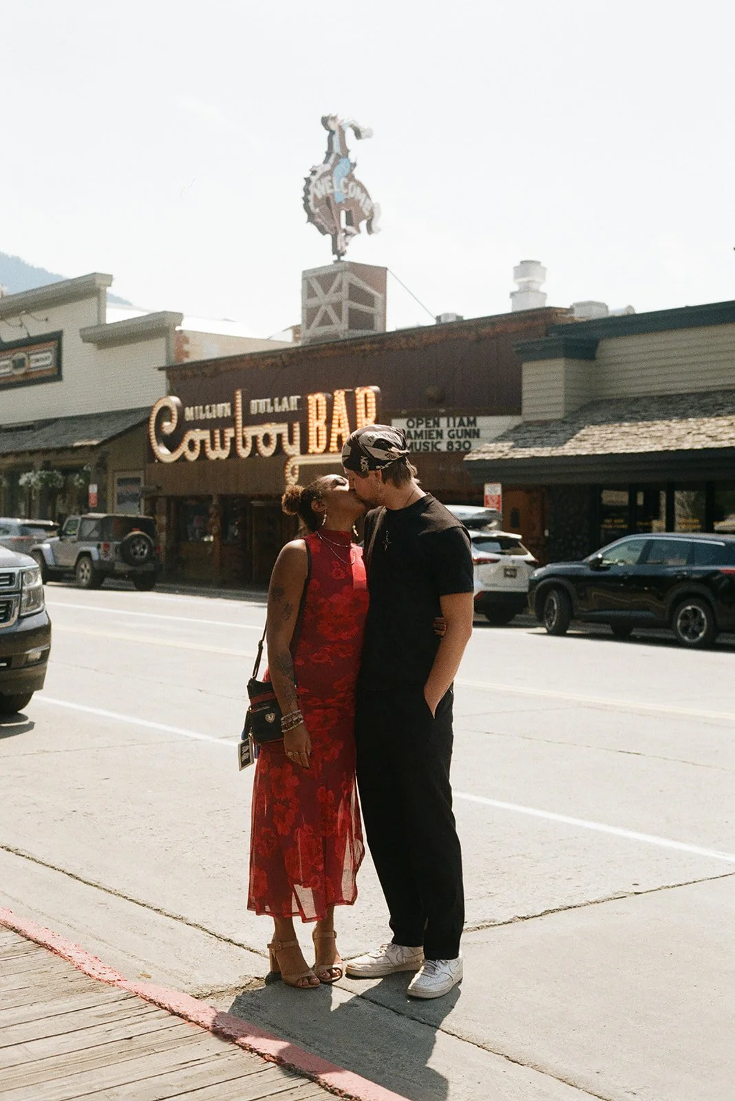 A couple kissing on a sidewalk in front of a building with a neon sign that reads "Million Dollar Courthouse" and a billboard above it. downtown jackson wyoming engagement shoot