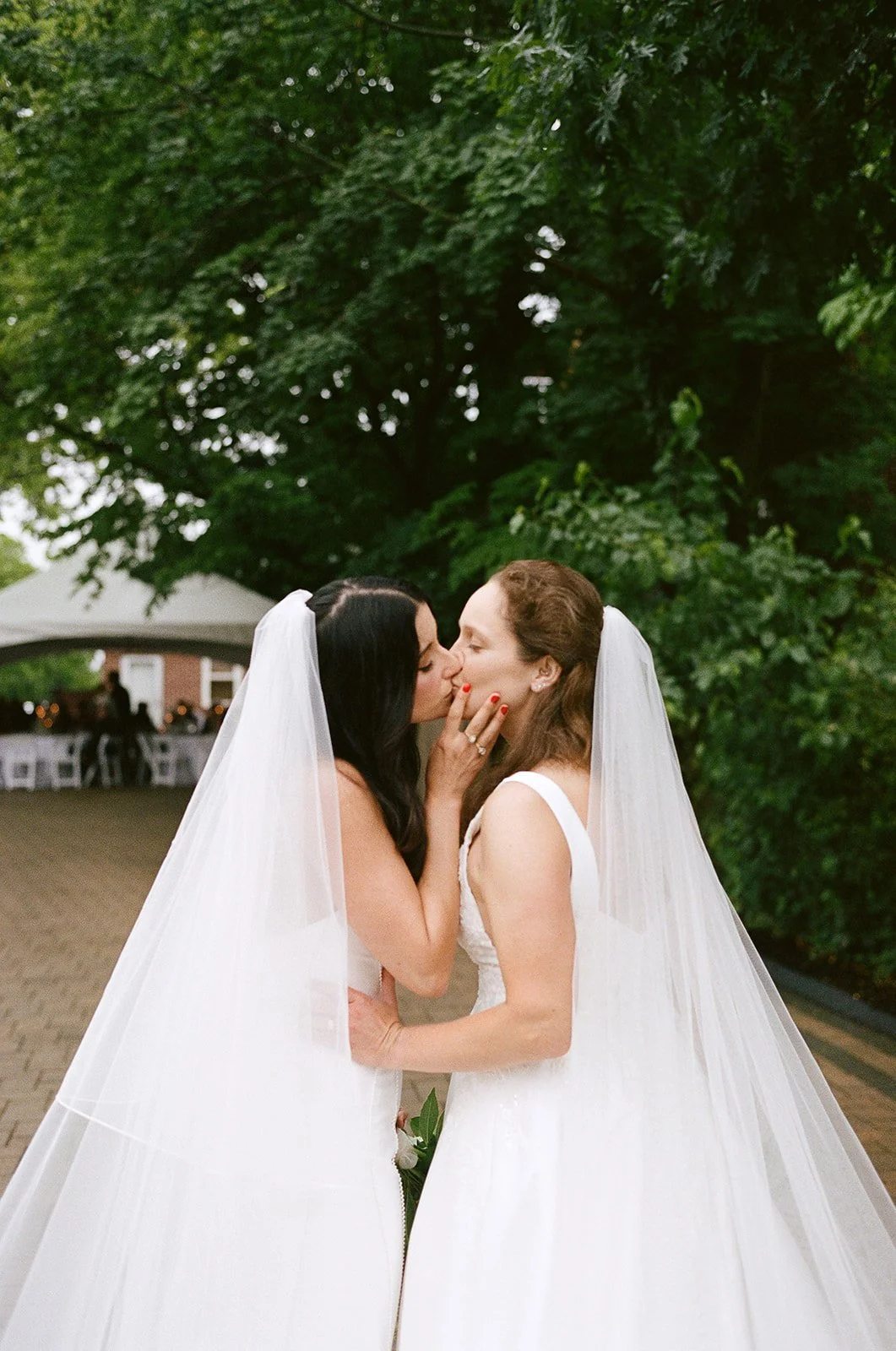 Two brides in wedding dresses and veils share a kiss outdoors surrounded by greenery. roanoke wedding film photographer virginia