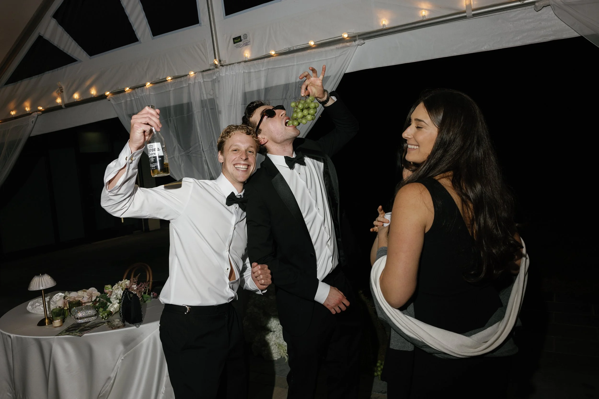 Three people dressed in formal attire at a celebration, with two men in tuxedos and a woman in a dress, standing under a tent at night. One man is holding a bunch of green grapes and bringing it to his mouth, another man is holding a beer bottle and smiling, while the woman is smiling and looking at the man with grapes. There is a table with floral arrangements and decorations in the background.