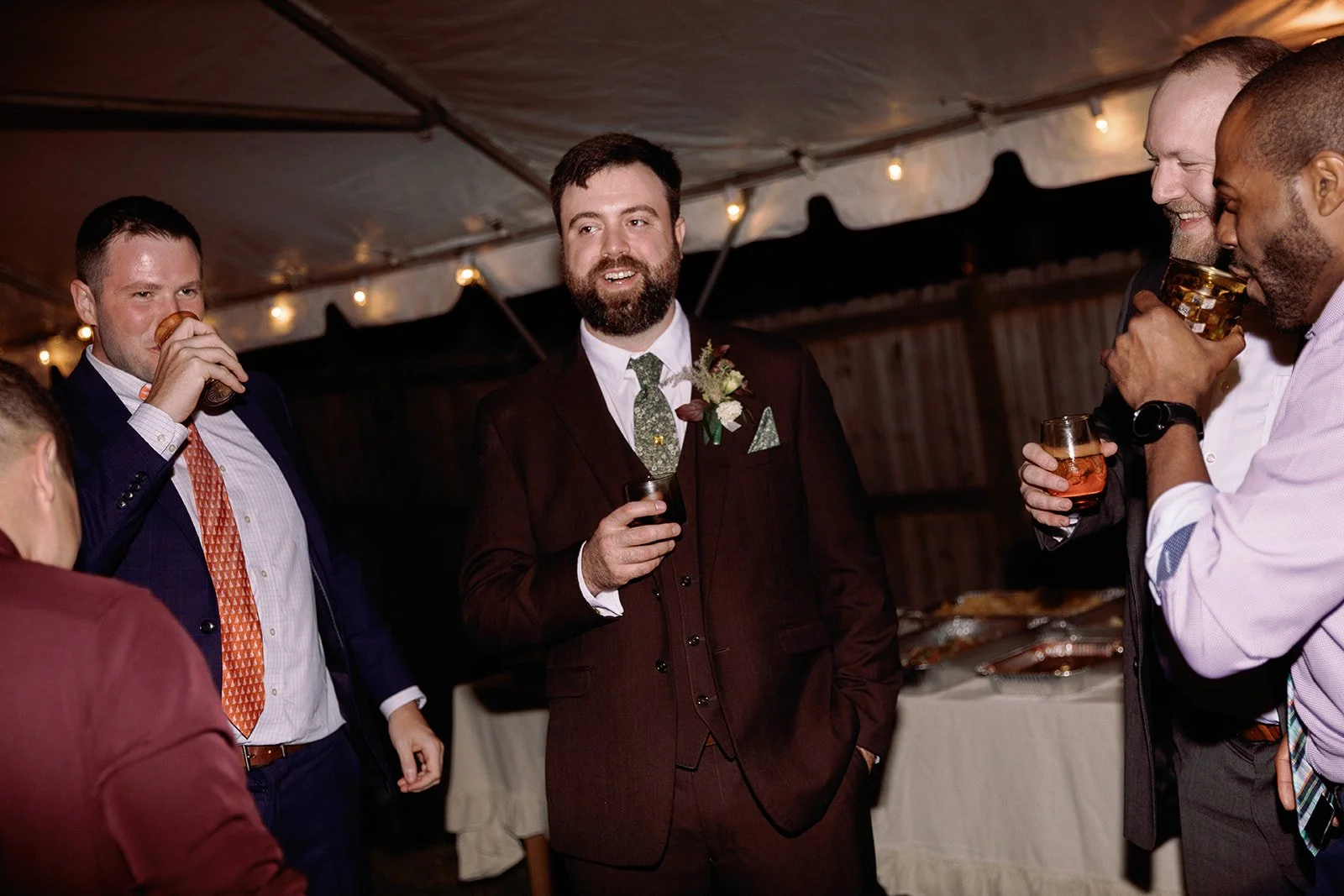 Men in suits and ties socializing and drinking at a gathering or celebration under a decorated tent. Staunton Virginia Wedding Film Photographer.