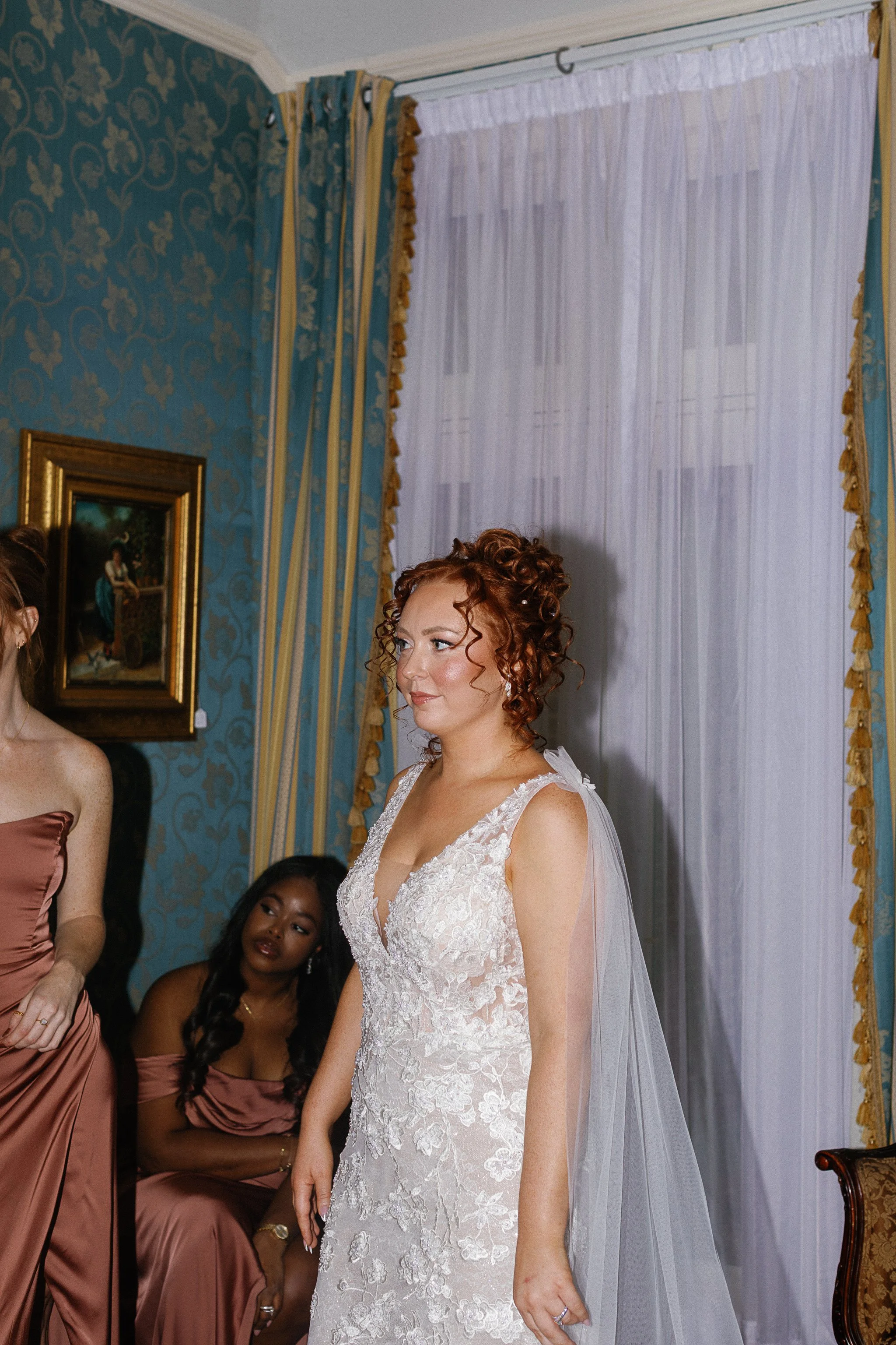 A bride in a lace wedding dress with red curly hair stands in a room with elegant patterned wallpaper and long curtains. A bridesmaid in a copper dress is partially visible beside her, and another seated bridesmaid in the background.