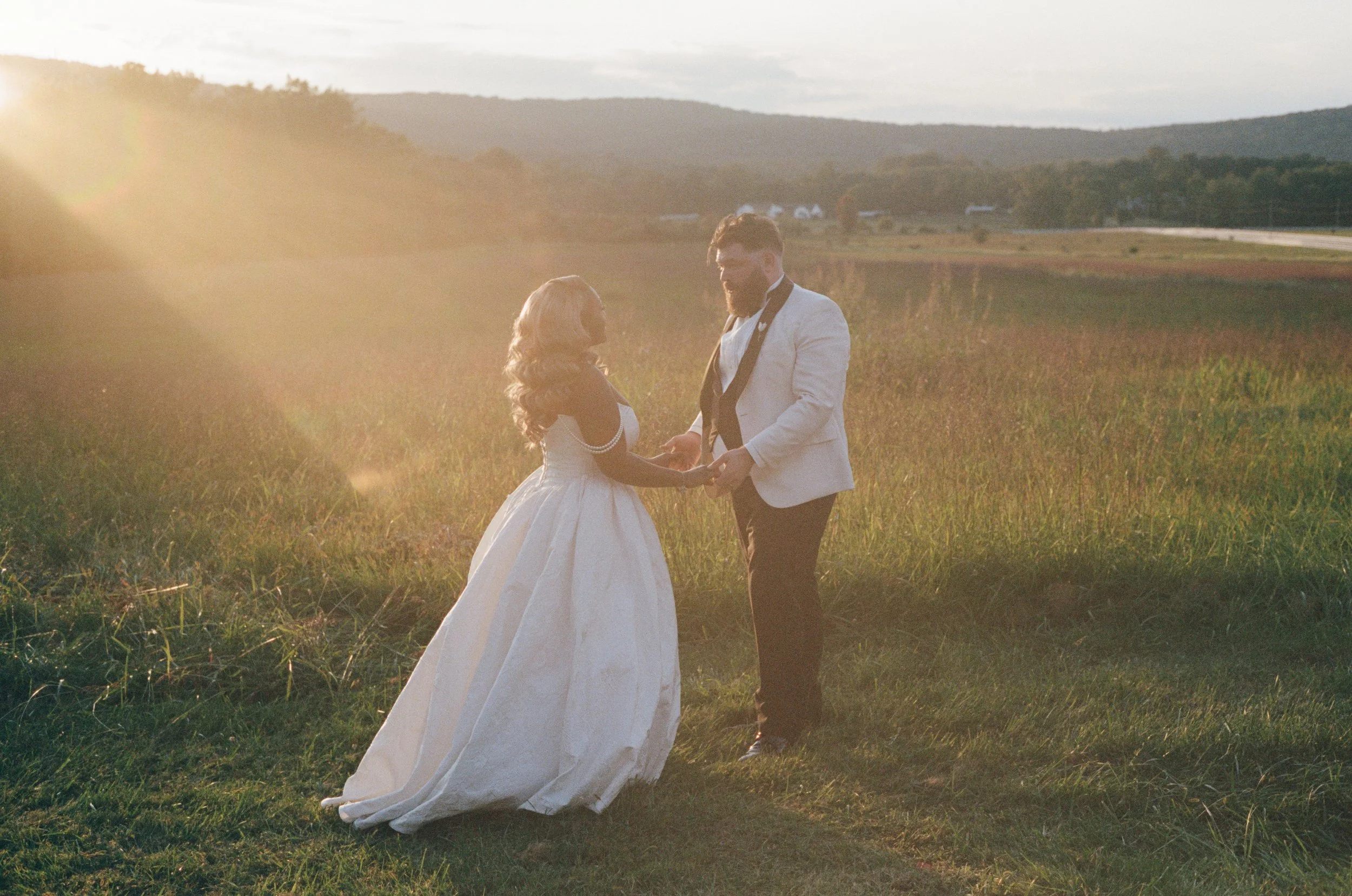 A couple in wedding attire holding hands in a grassy field during sunset, with mountains and a few distant buildings in the background at High Torr Estate in Purcellville Virginia.