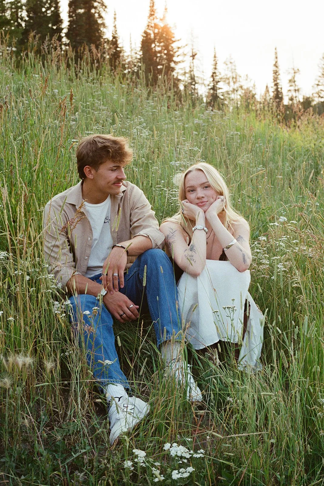 A young man and woman sitting in a grassy field at sunset, with a forest in the background. The woman rests her chin on her hands, and the man looks at her, both appearing relaxed and contemplative. bozeman montana photographer