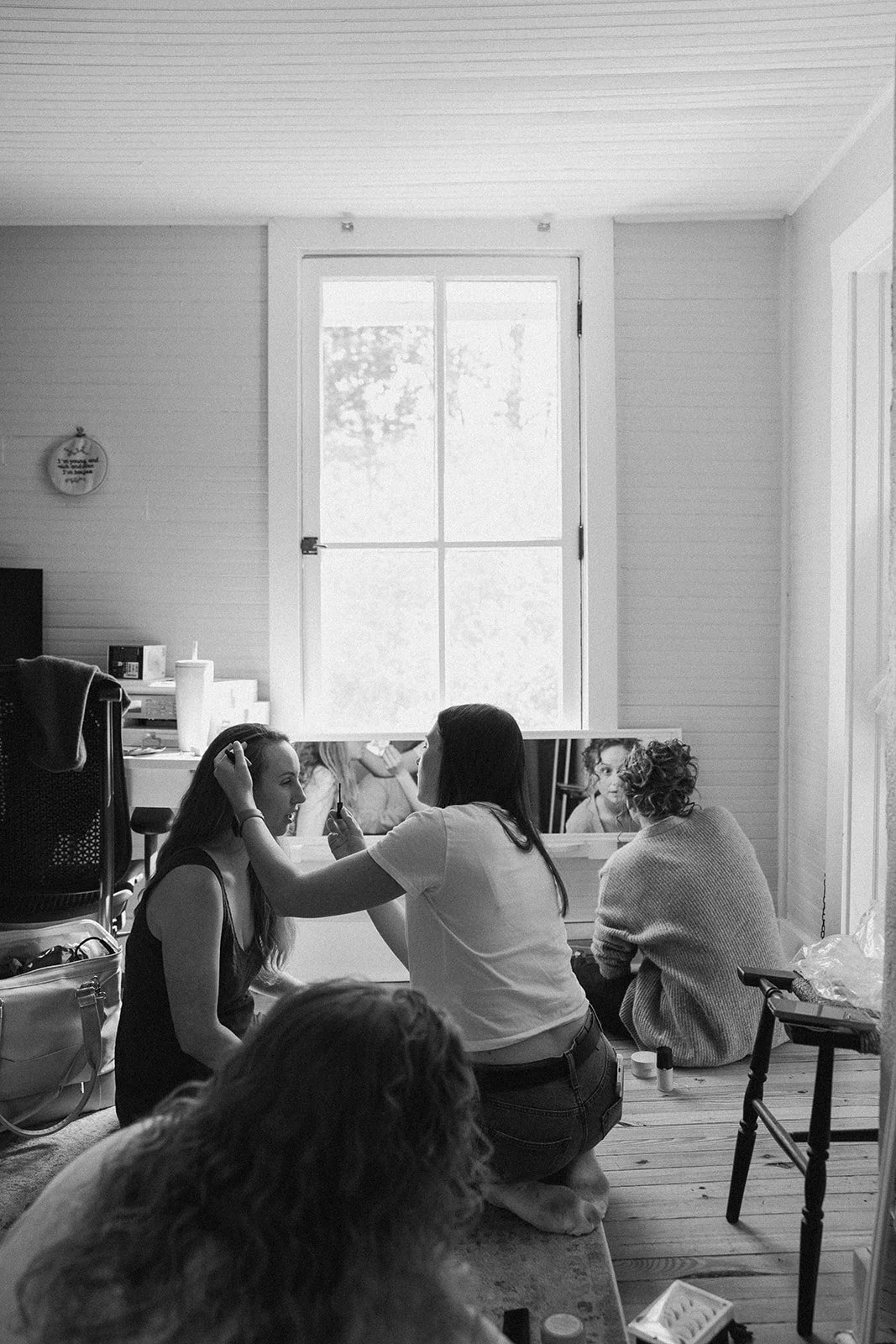 Women gathered in a room, some applying makeup, with a window in the background. charlottesville virginia Wedding Film Photographer. backyard wedding
