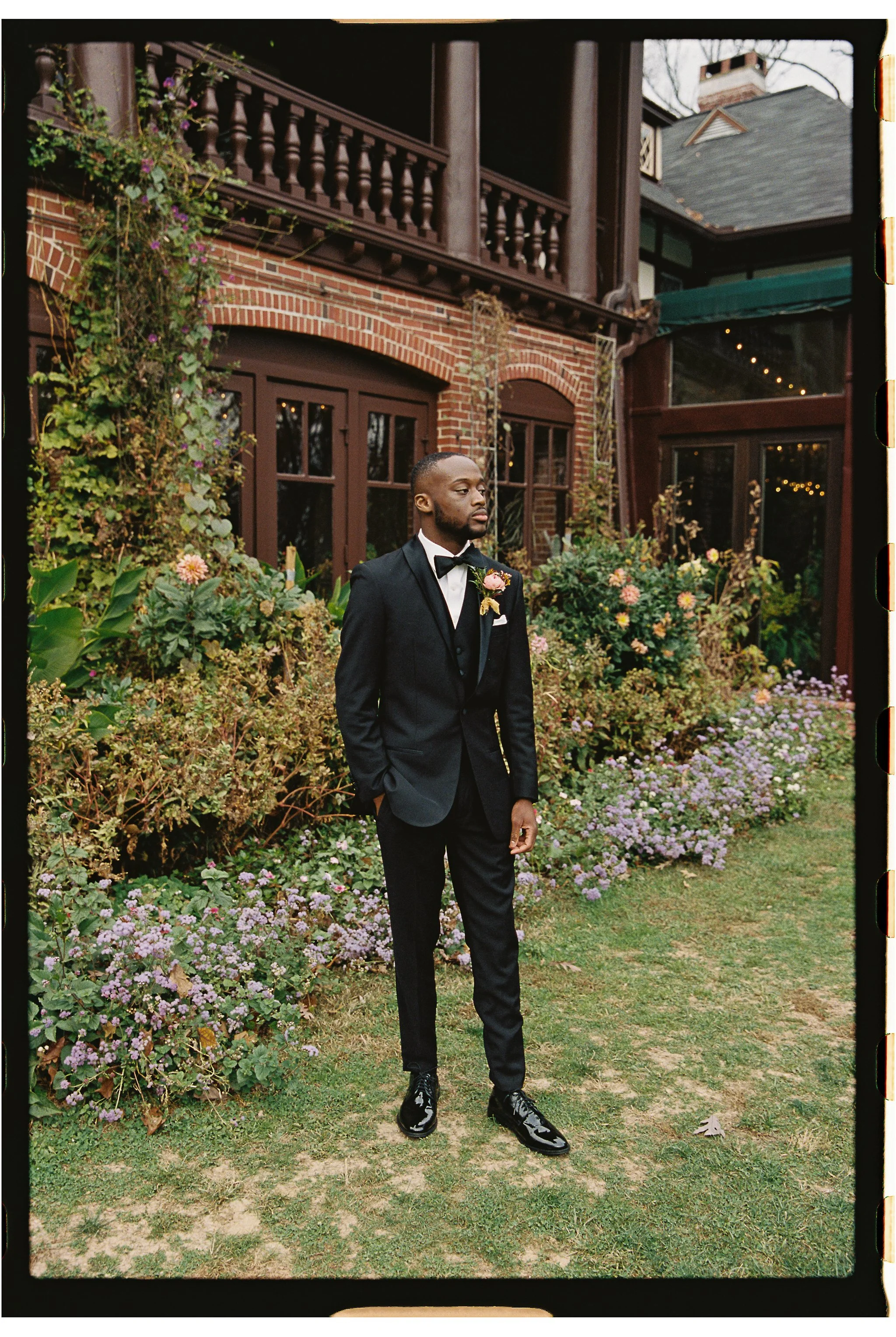 A man in a formal black suit and bow tie standing on a lawn in front of a brick building with a garden of purple flowers and greenery. gramercy mansion maryland wedding