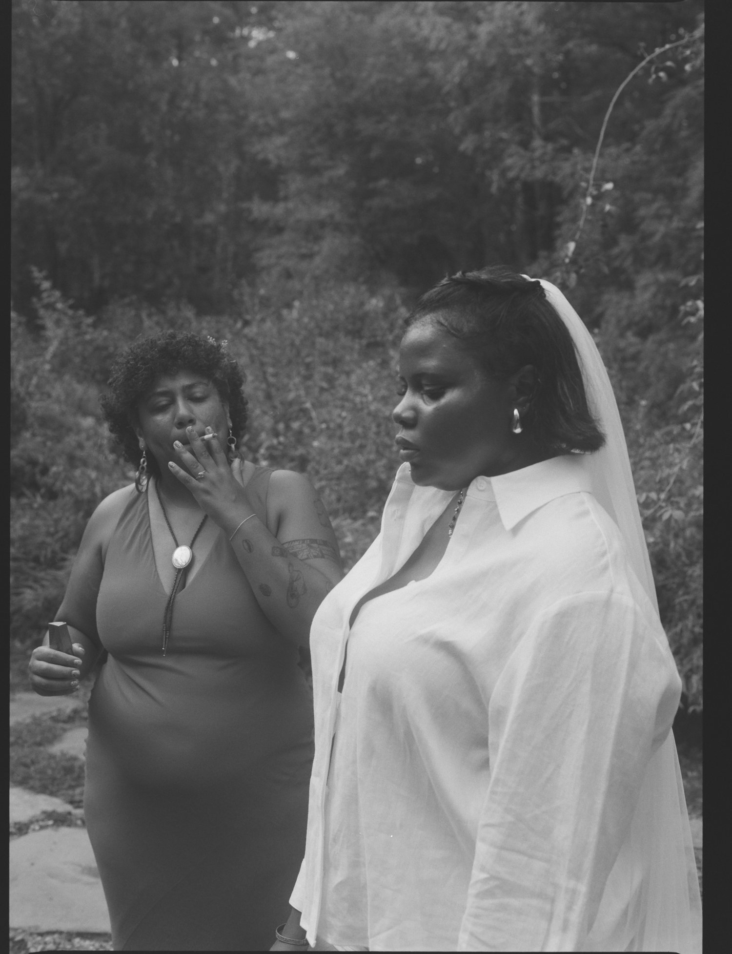 Queer couple outdoors, one smoking a cigarette and the other wearing a veil, in a black and white photo.