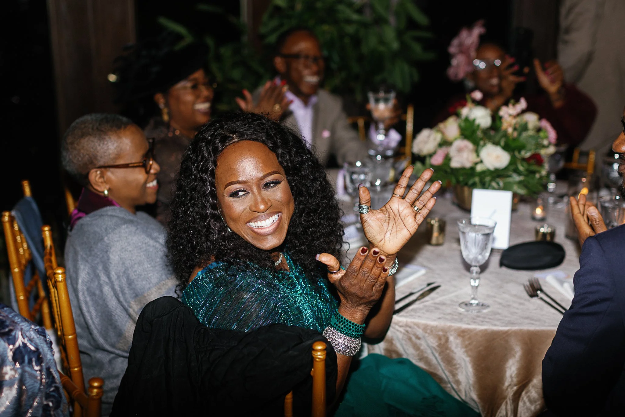 A joyous woman clapping and smiling at a celebratory event with a group of people seated around a table adorned with a floral arrangement. gramercy mansion maryland wedding