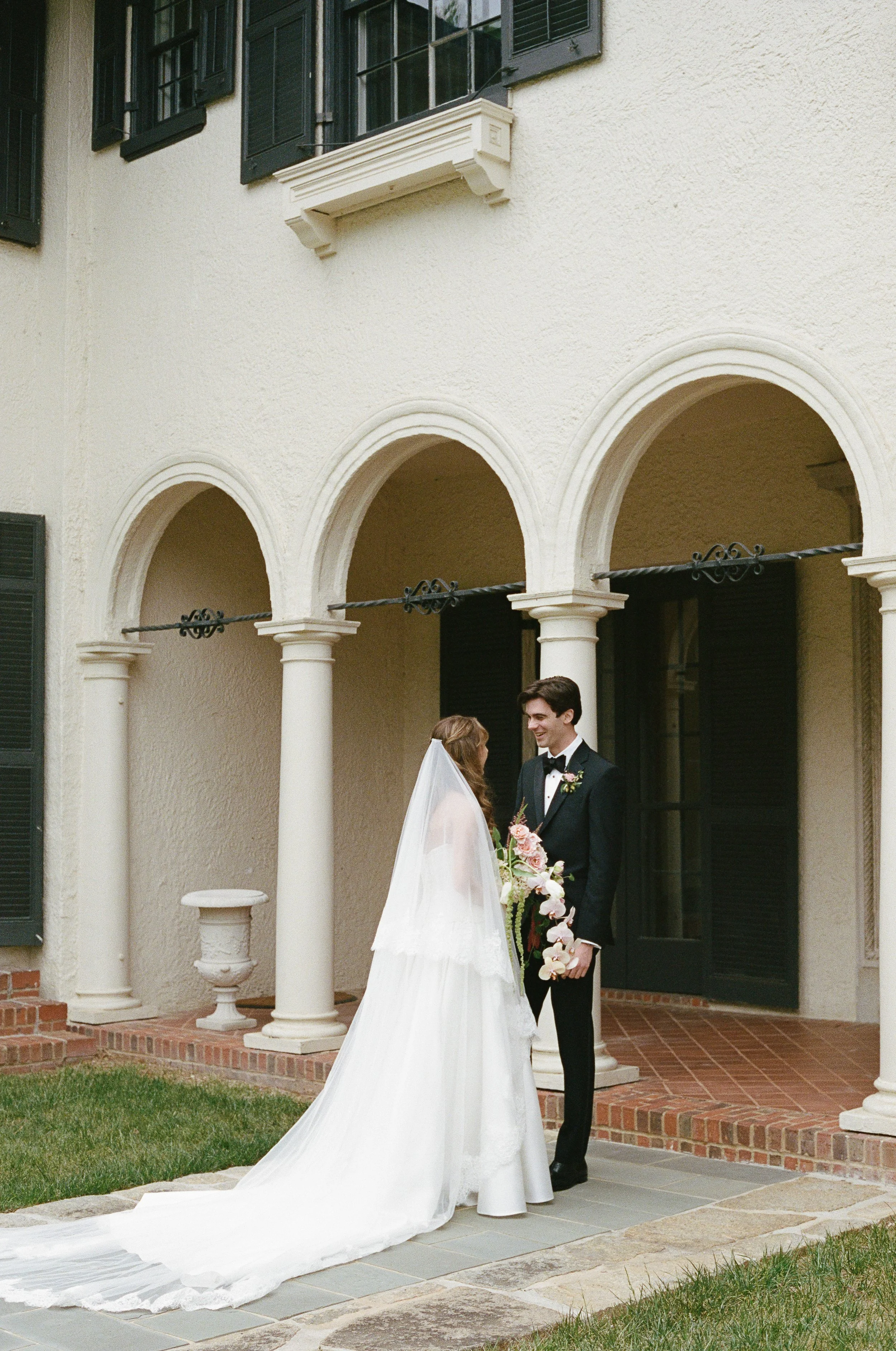 A bride and groom are standing outside a building with white walls and black shutters, under arched columns. The bride is wearing a white wedding dress with a long train, and the groom is in a black tuxedo. They are holding hands and smiling at each other, with the bride holding a bouquet of pink and white flowers.