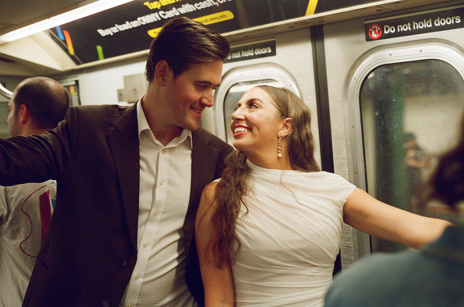 A man and woman on a subway train smiling at each other, standing close near the door, with the woman reaching towards the window. Brooklyn New York Wedding Film Photographer