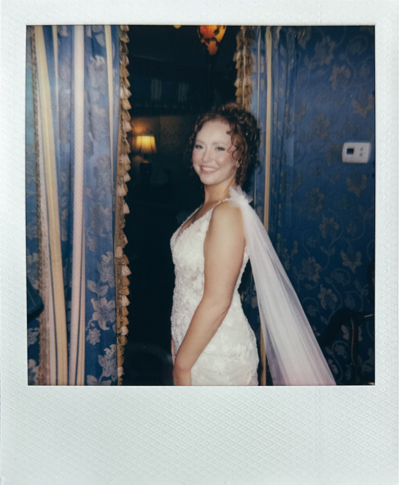 A woman in a white wedding dress with lace details and a veil, smiling, standing inside a room with blue floral wallpaper, curtains, and a lamp in the background. Gramercy Mansion Wedding Film Photographer.