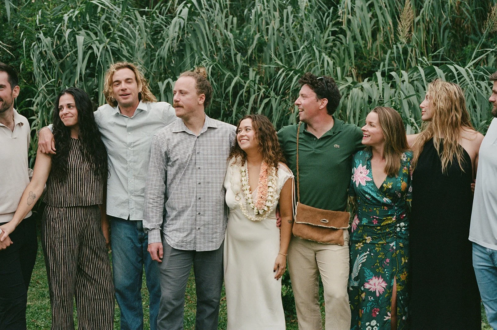 Group of people standing together outdoors near tall green plants, smiling and hugging each other. ocracoke outerbanks wedding film photographer