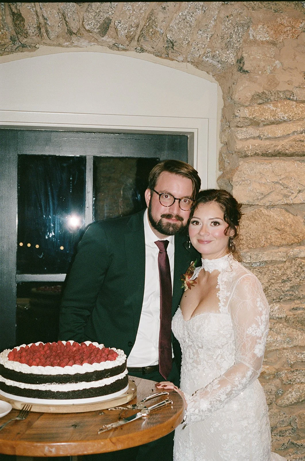 A bride and groom standing together indoors near a window with a stone wall background, with a wedding cake on a table in front of them. potters craft cider charlottesville va