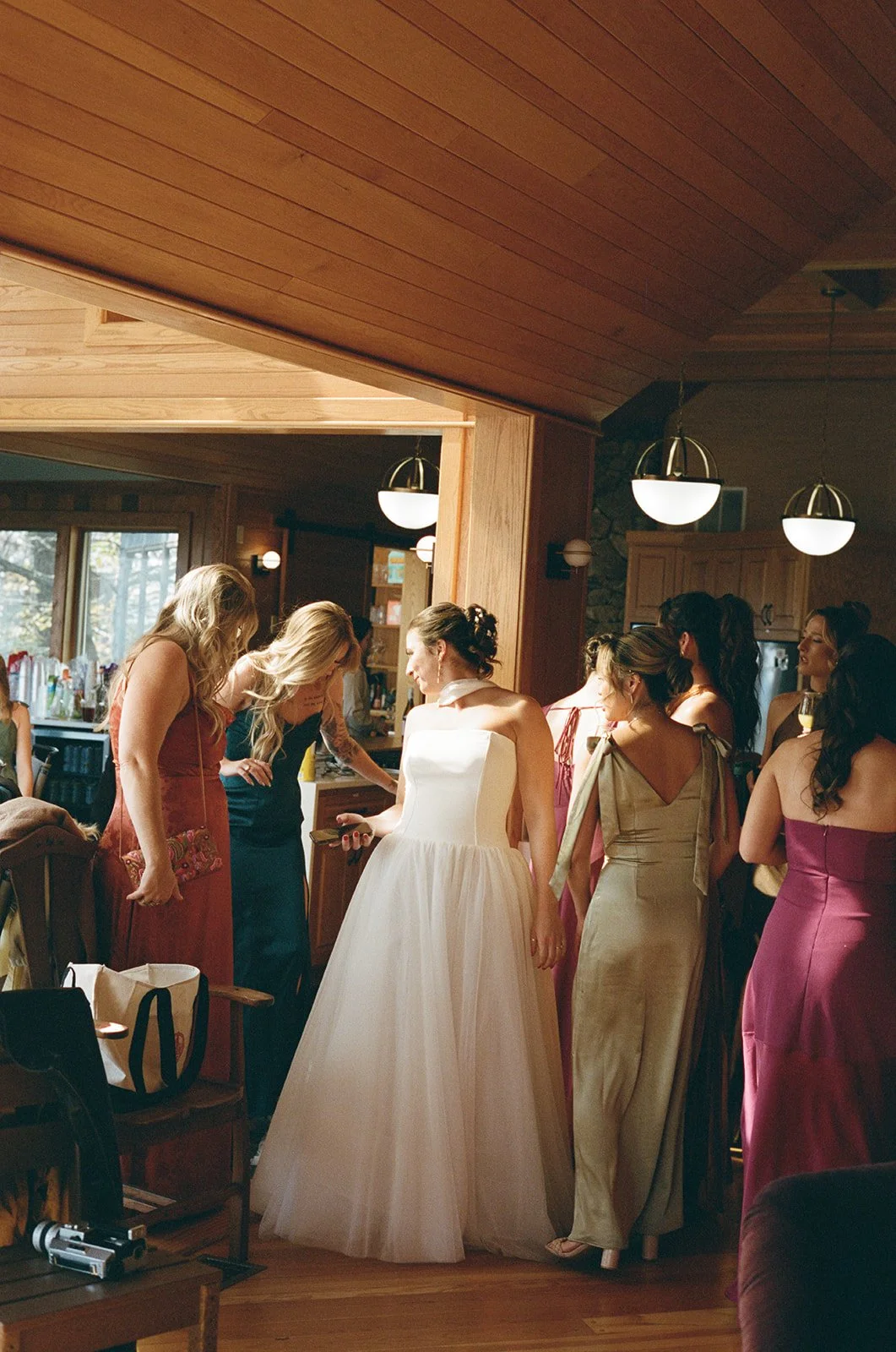 A group of women, including a bride in a white wedding dress, gathered in a warmly lit wooden kitchen or dining area. Wintergreen Resort Virginia Wedding Film Photographer