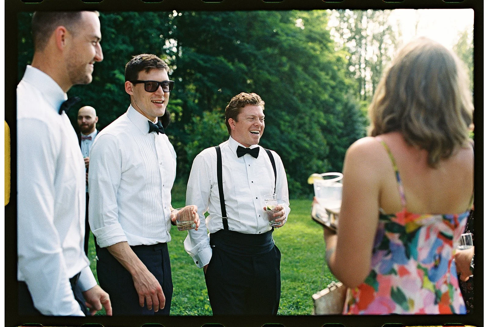Group of men in tuxedos and a woman in a floral dress having a conversation outdoors during a gathering or party. Charlottesville Virginia Wedding Film Photographer