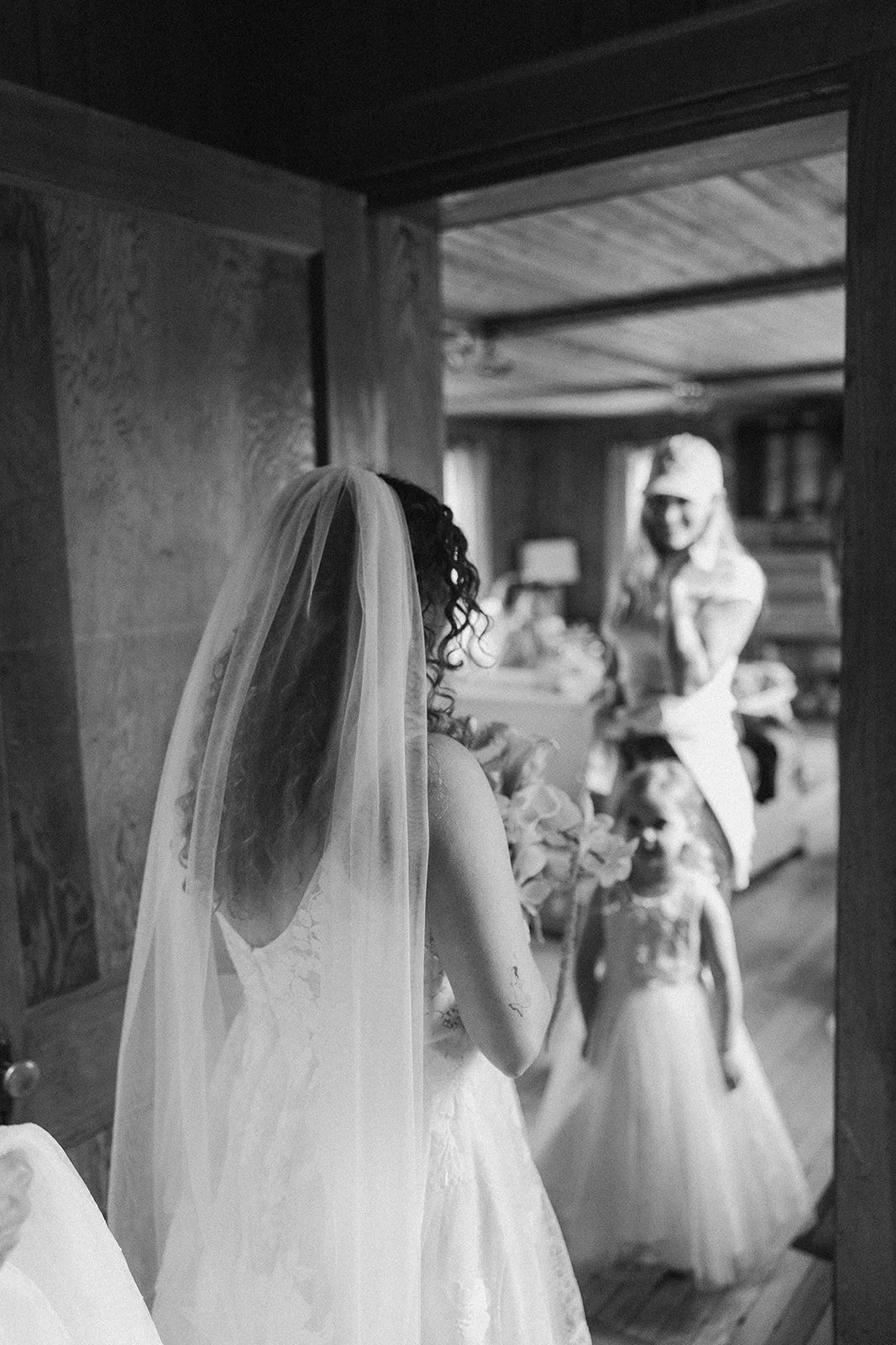 A black and white photo of a bride with curly hair, wearing a wedding dress and veil, standing and holding flowers. In the background, two young girls in dresses and a woman in a white outfit and hat are visible in a cozy indoor setting.