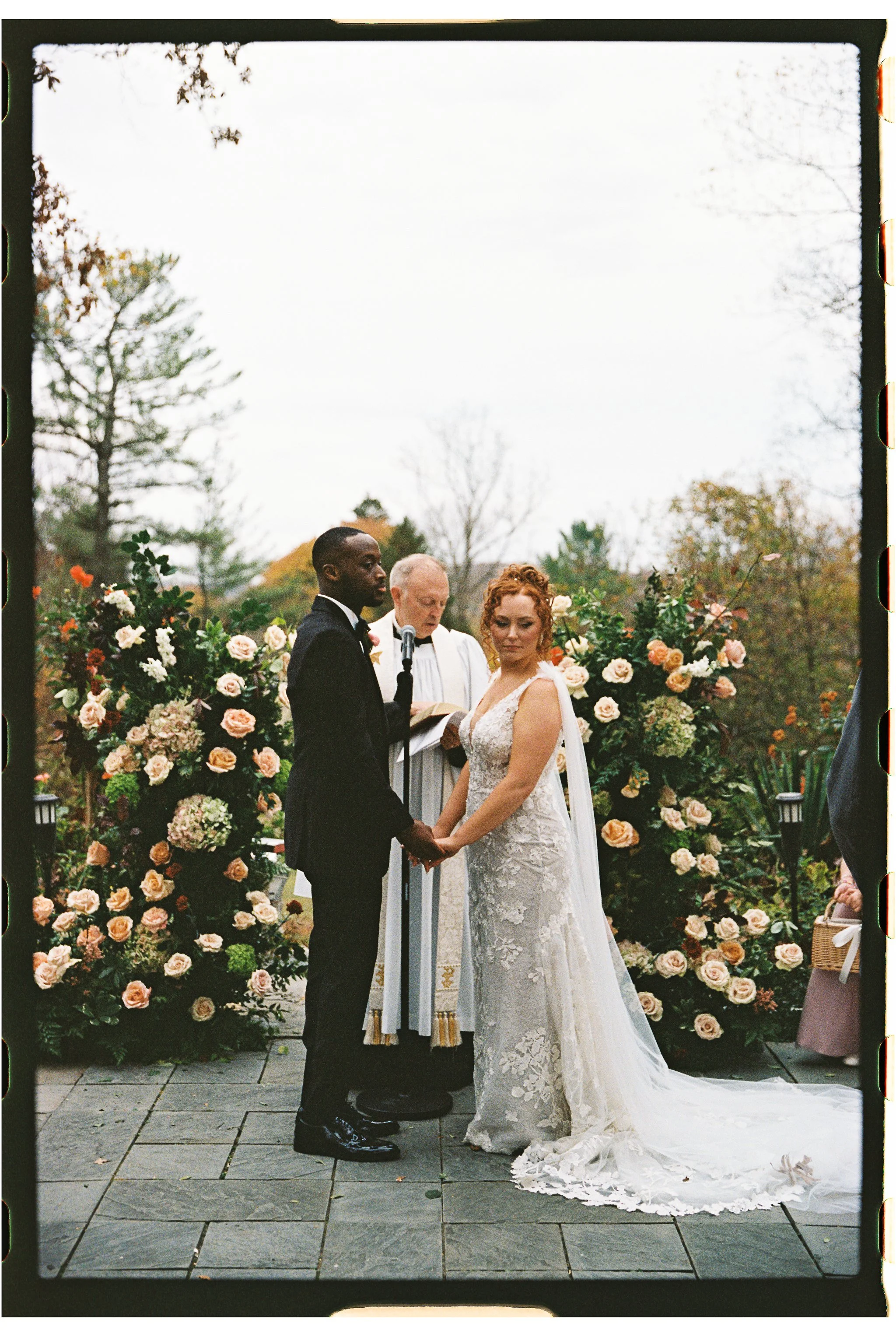 A couple holding hands during an outdoor wedding ceremony, with a priest officiating. The bride is wearing a lace wedding gown and the groom is in a tuxedo. There are floral arrangements in the background. gramercy mansion maryland wedding