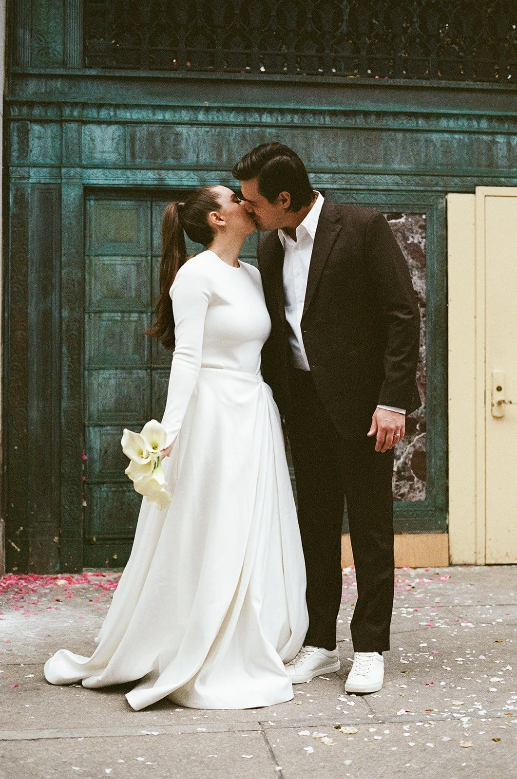 A bride and groom share a kiss outside, with pink petals scattered on the ground. The bride is wearing a white dress and holding a bouquet of white calla lilies, while the groom is dressed in a black suit and white sneakers. They are standing in fron