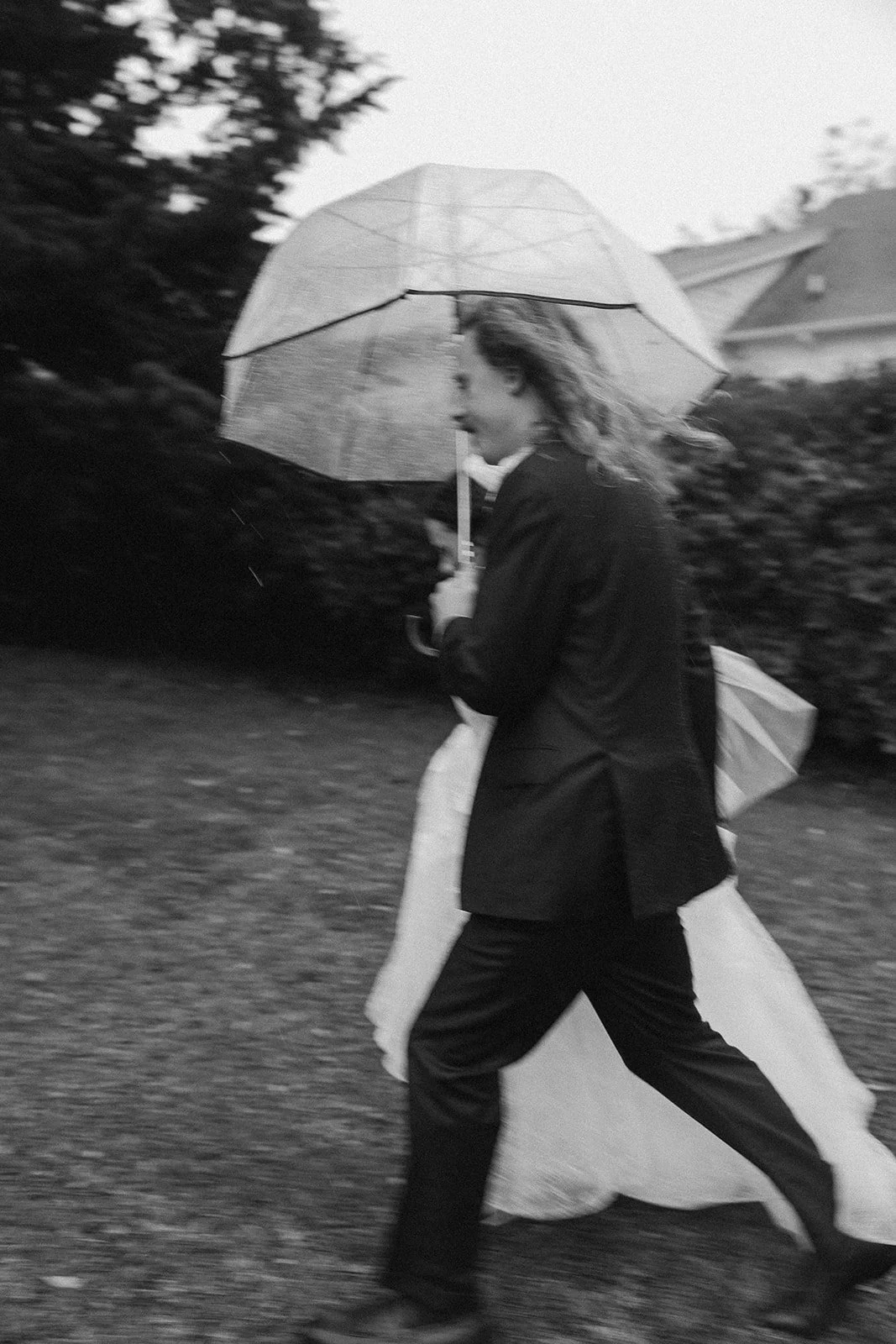 A woman walking outdoors in the rain, holding an umbrella. She is dressed in formal attire with a black coat and appears to be carrying a large bag or possibly a wedding dress. ocracoke outerbanks wedding film photographer