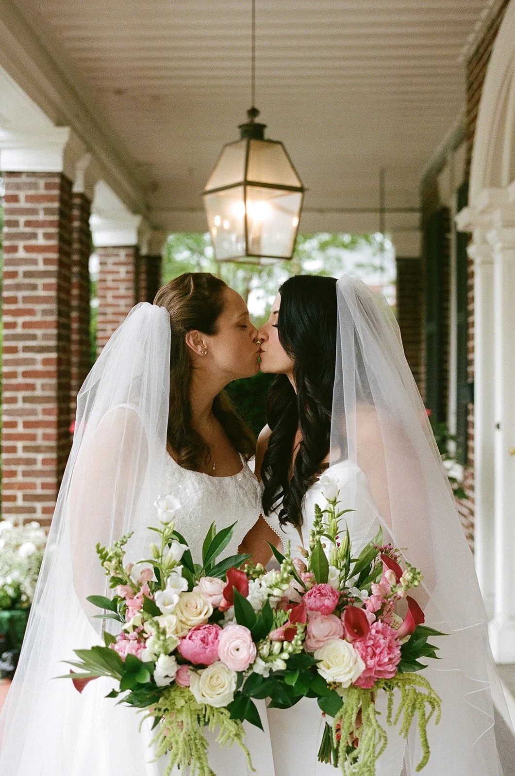 Two brides in wedding dresses and veils kiss on a porch, holding a large bouquet of pink and white flowers. roanoke wedding film photographer virginia