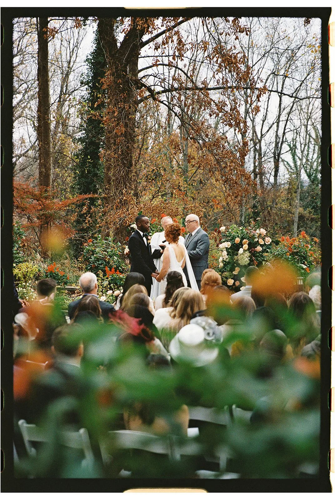 A wedding ceremony taking place outdoors in a wooded area with tall trees and fall foliage, with the bride and groom holding hands and facing each other, surrounded by a group of guests, and officiated by a person in a gray suit. Gramercy Mansion.