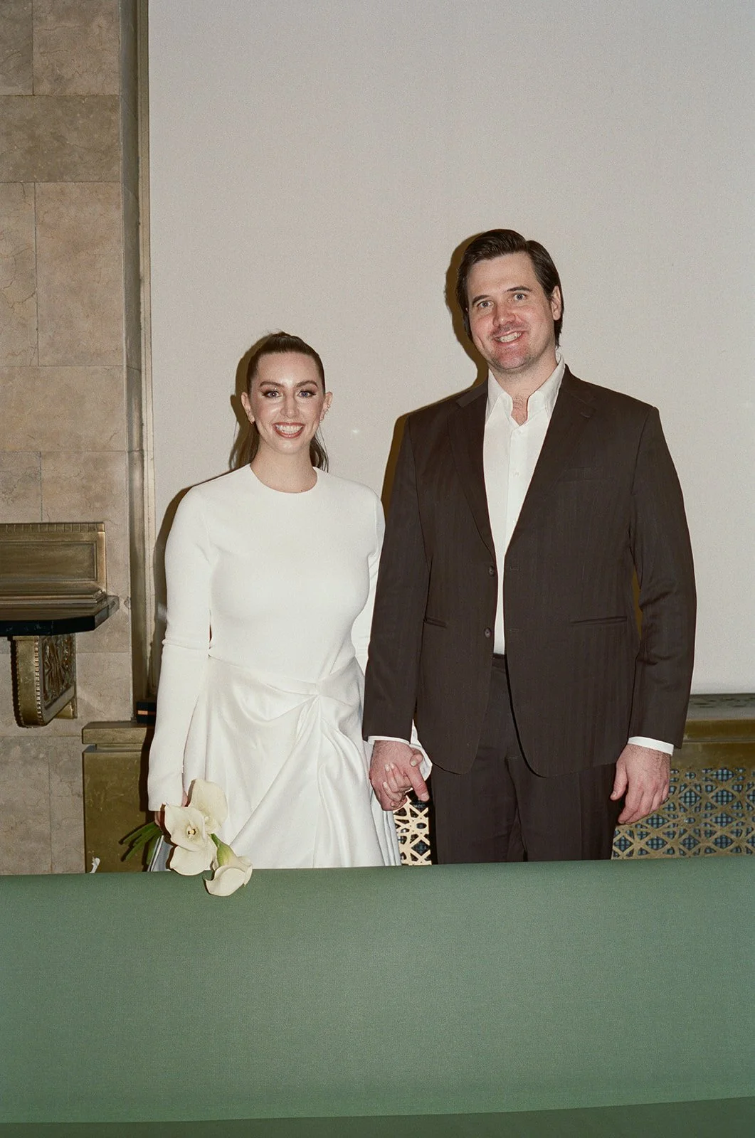 A bride and groom holding hands at their wedding, standing behind a green table with a white flower on it, in an indoor setting. NYC city hall elopement wedding film photographer