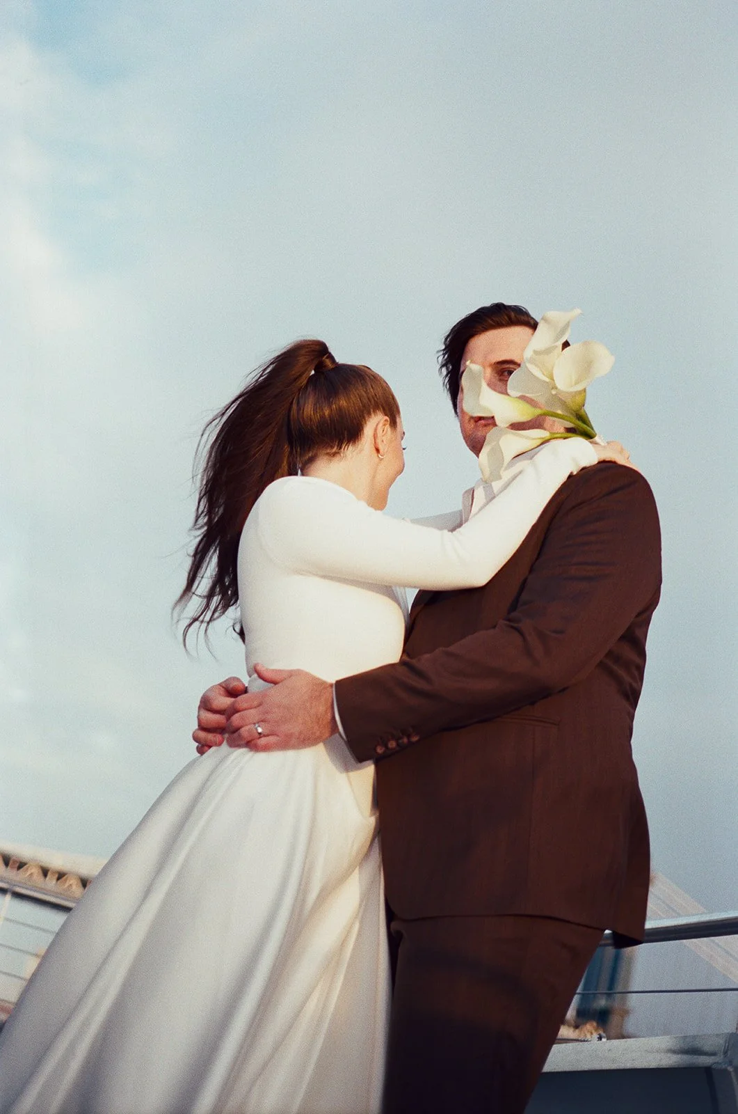 A woman in a white dress and a man in a brown suit embrace outdoors with a bouquet of white flowers, against a blue sky background. New York City Elopement Wedding Film Photographer