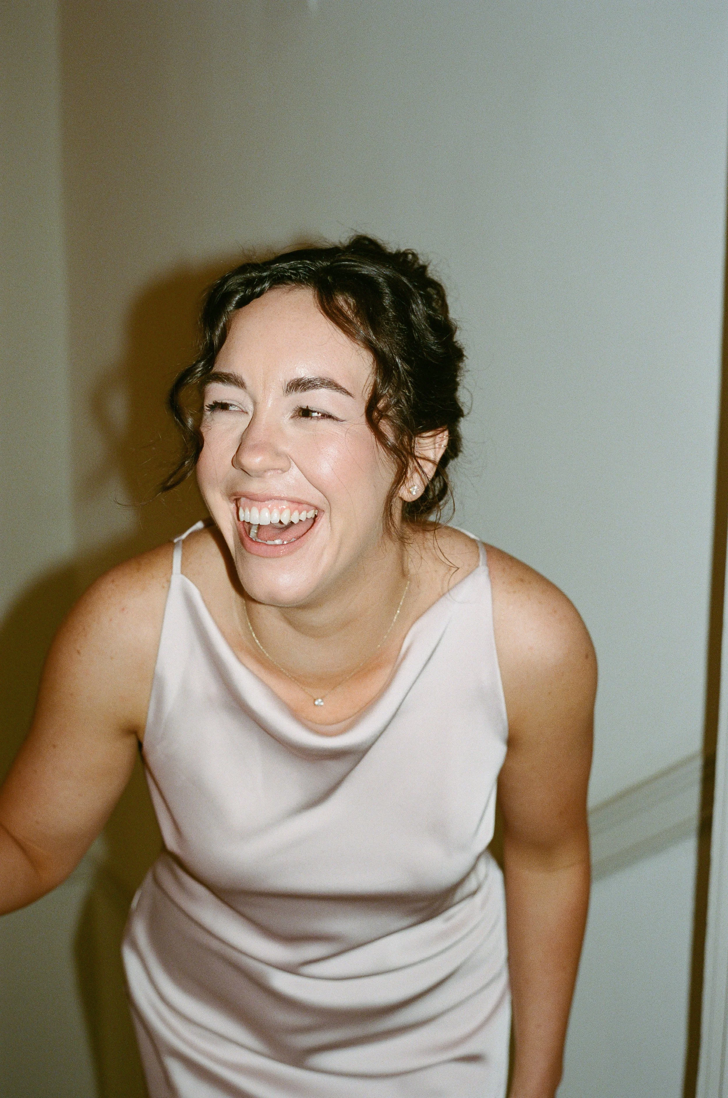 A woman with dark, curly hair laughing, wearing a white sleeveless dress and a delicate necklace, standing against a plain wall.