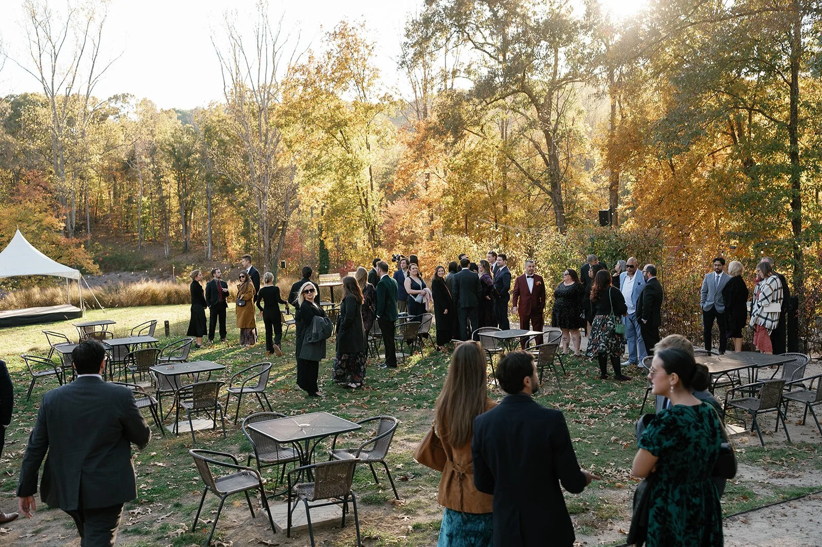 Group of people dressed in formal attire outdoors in a park with trees showing fall foliage, some standing and chatting, some seated at tables. potters craft cider charlottesville va