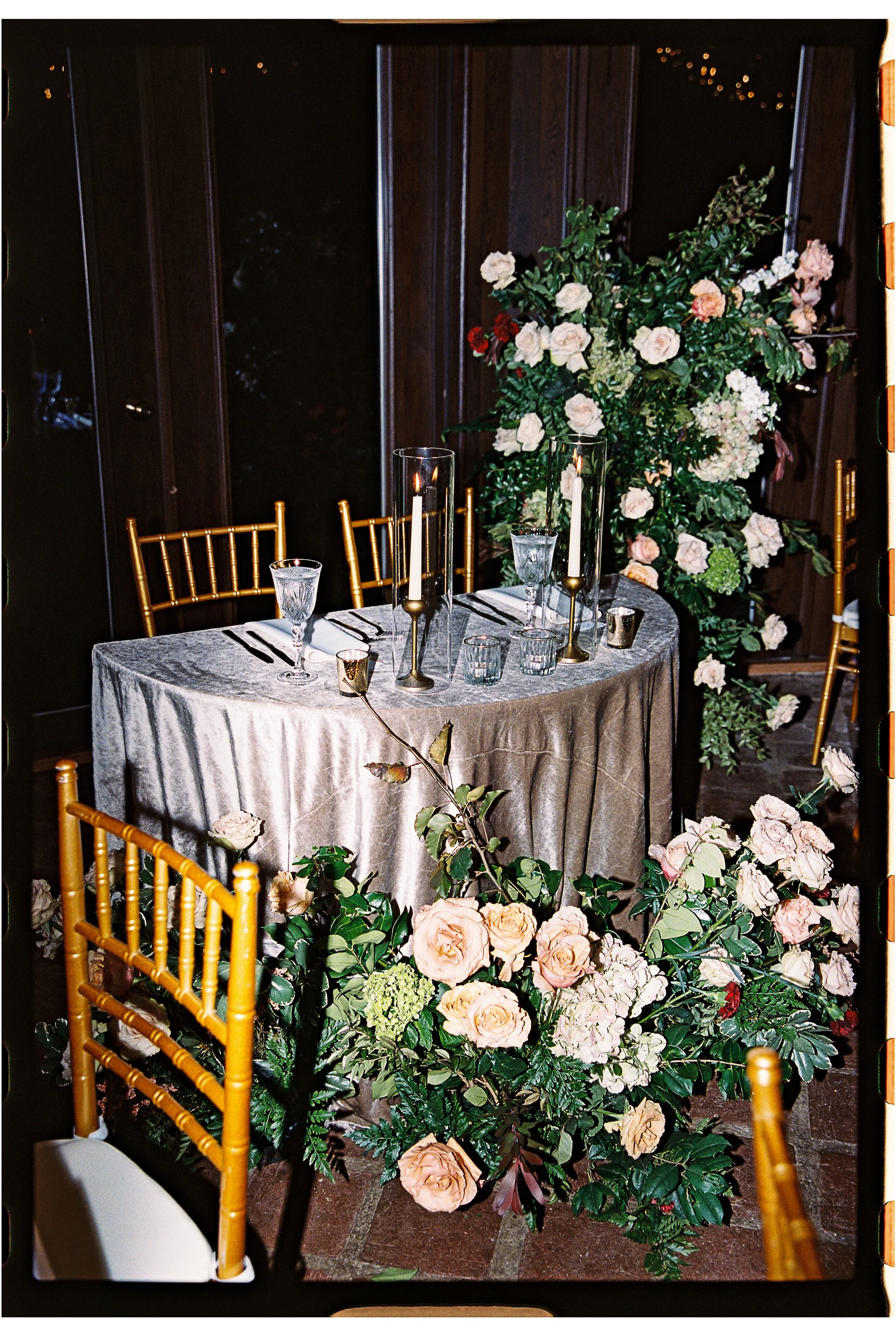 Elegant table setting with velvet tablecloth, glassware, candlesticks, and gold chairs, surrounded by lush floral arrangements with roses and greenery. gramercy mansion maryland wedding 35mm film