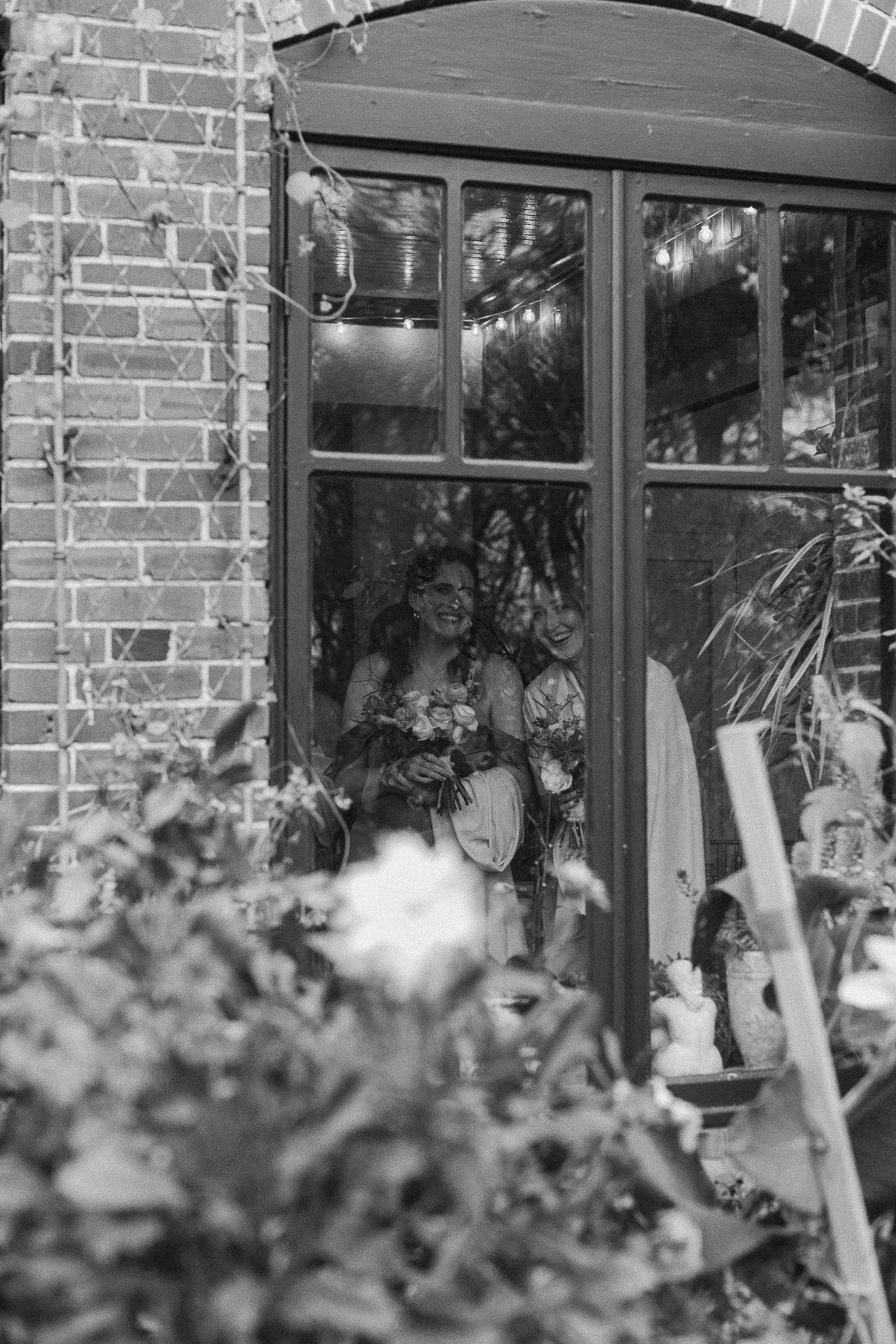 Black and white photo of two people smiling and holding bouquets inside a building seen through a window, surrounded by brick and foliage. gramercy mansion maryland wedding