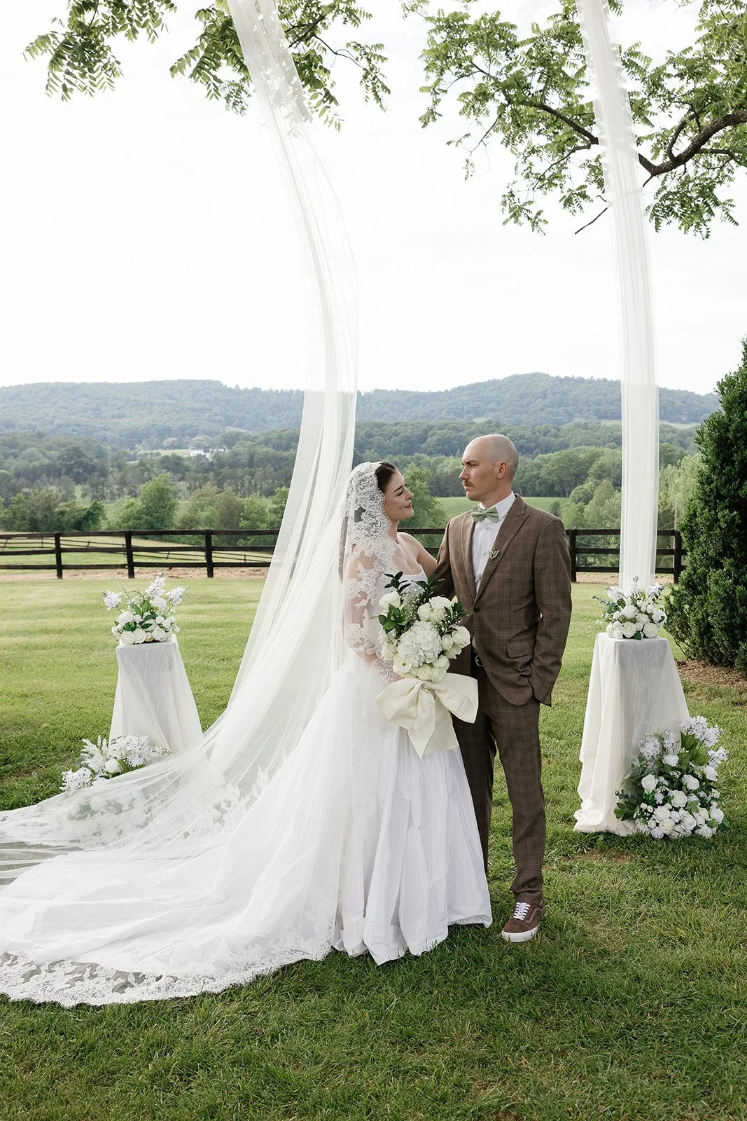 A bride and groom standing outdoors during their wedding ceremony, with the bride holding a bouquet of white flowers and wearing a lace wedding dress and veil, and the groom wearing a brown plaid suit with a light-colored bow tie. There are white flower arrangements on pedestals on either side of them, and a scenic landscape with green hills and a wooden fence in the background.
