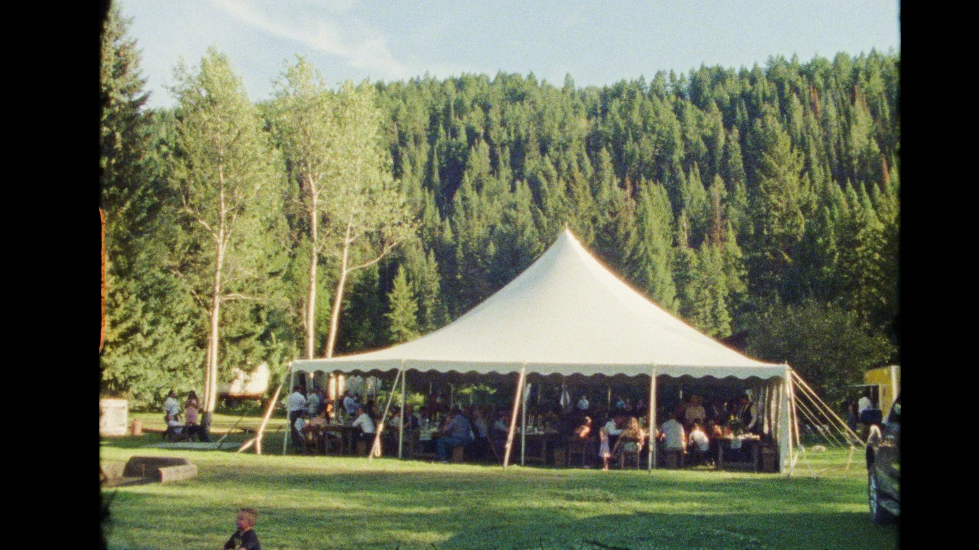 A large white event tent set up outdoors in a park with green grass and tall trees in the background, and people sitting at tables underneath the tent. Moose Creek Ranch Super8