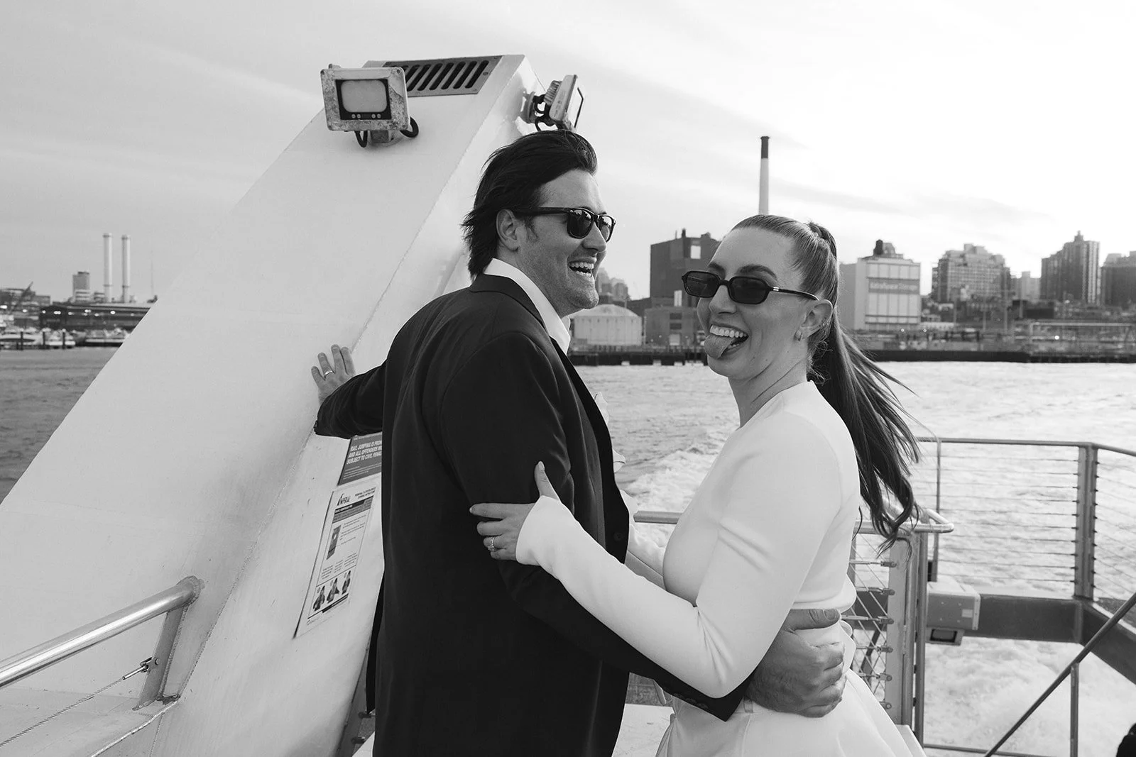 A black and white photo of a smiling couple, a man and a woman, on a boat with a city skyline in the background. They are wearing sunglasses and playful, sticking out their tongues, with the woman holding the man around his waist. NYC elopement Ferry