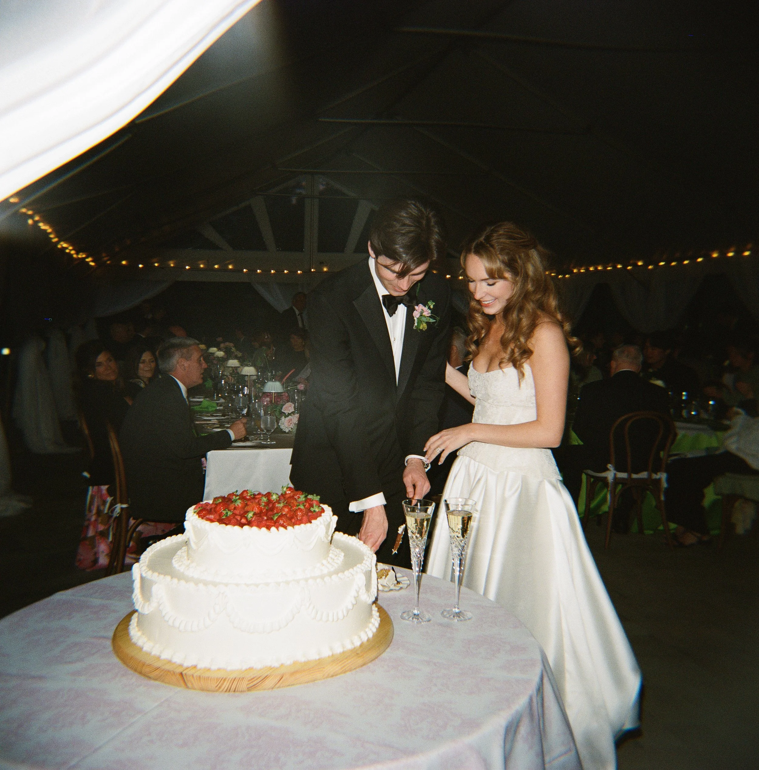 A wedding reception with a bride and groom cutting a wedding cake with strawberries on top, while guests are seated at tables in the background.