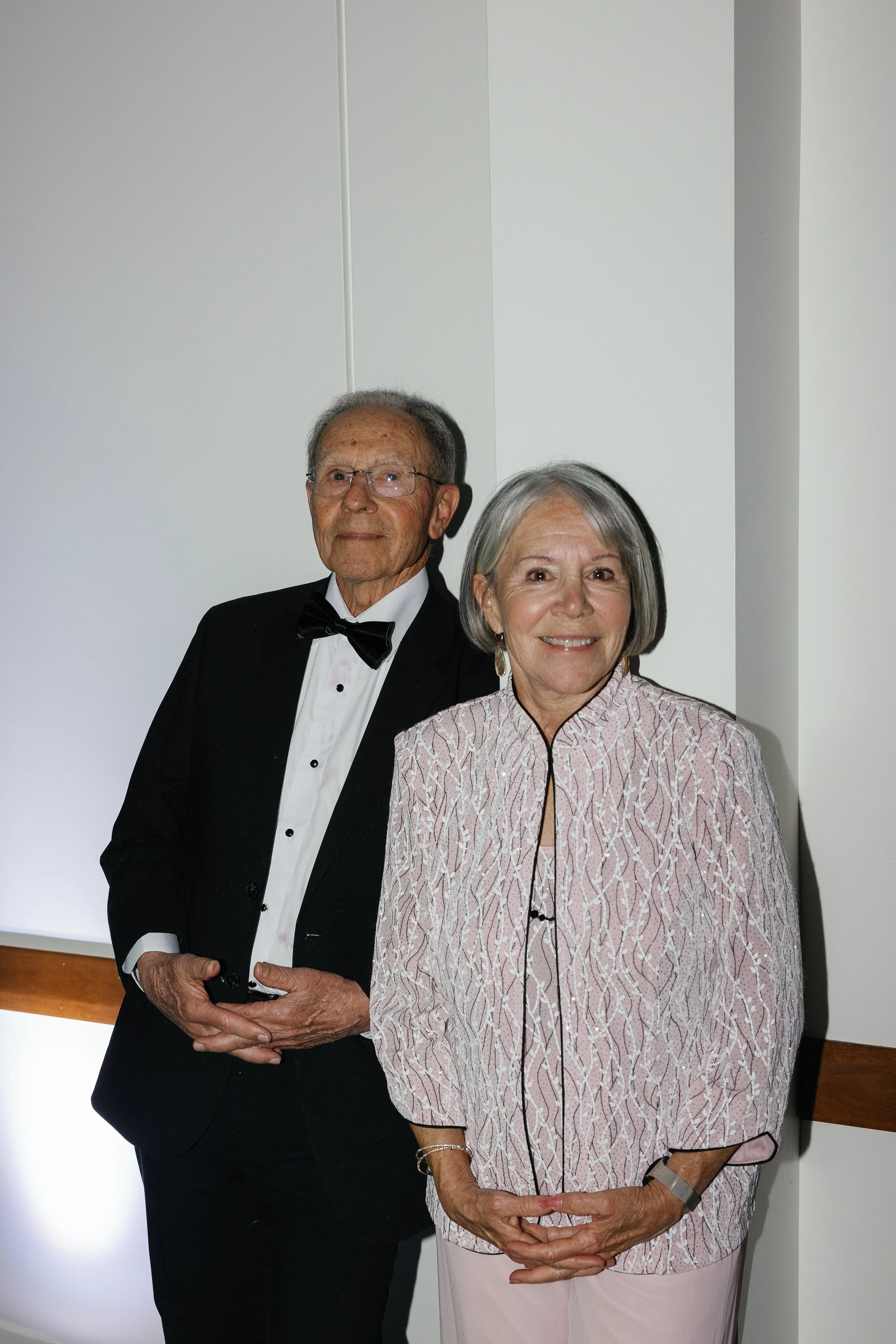 An elderly man in a tuxedo with a bow tie and an elderly woman in a matching patterned jacket and light-colored pants standing together in front of a plain wall, smiling at the camera. richmond virginia wedding photographer