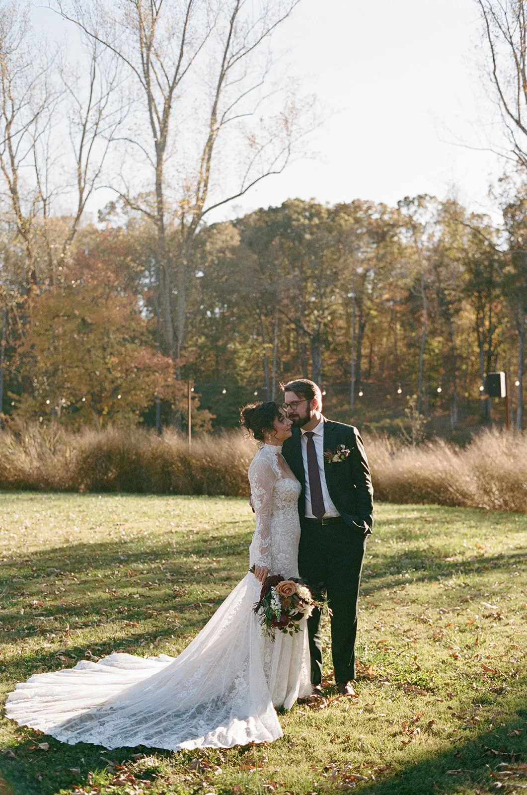 A bride and groom share an intimate moment outside on a grassy field with autumn trees in the background. The bride wears a lace wedding gown with a long train, and the groom wears a dark suit with a boutonnière. The bride holds a bouquet of flowers.