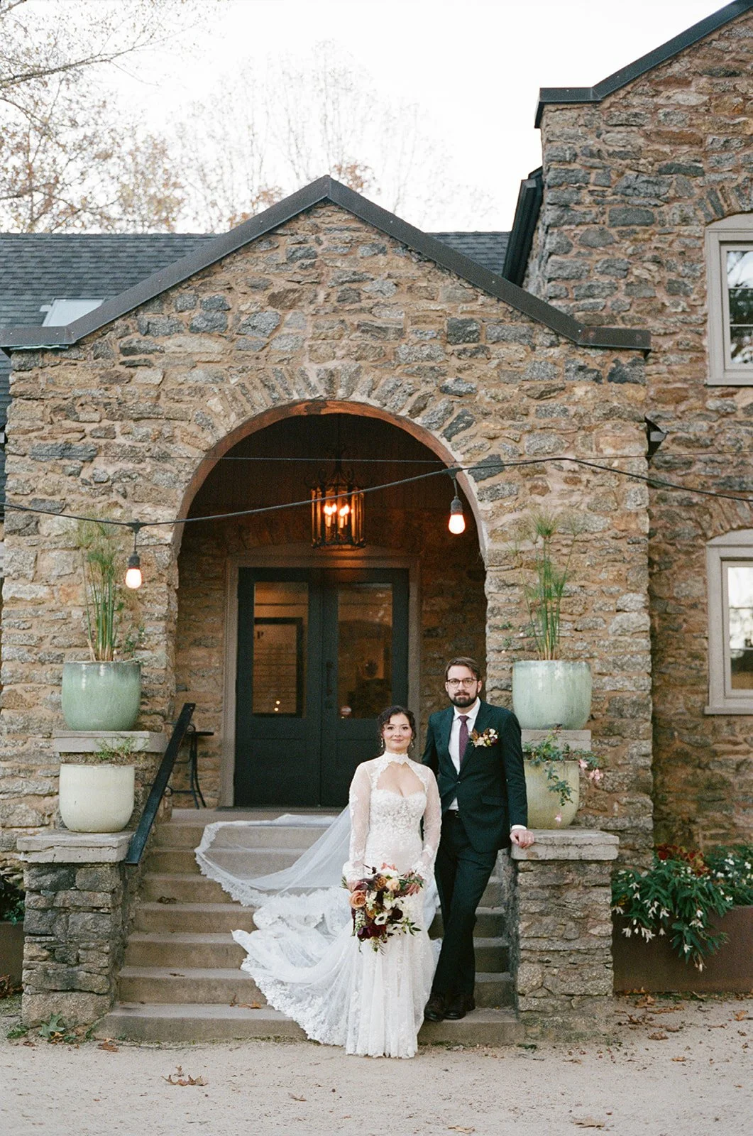 A bride and groom standing on stone steps in front of a rustic stone building with large potted plants and string lights overhead. potters craft cider charlottesville va
