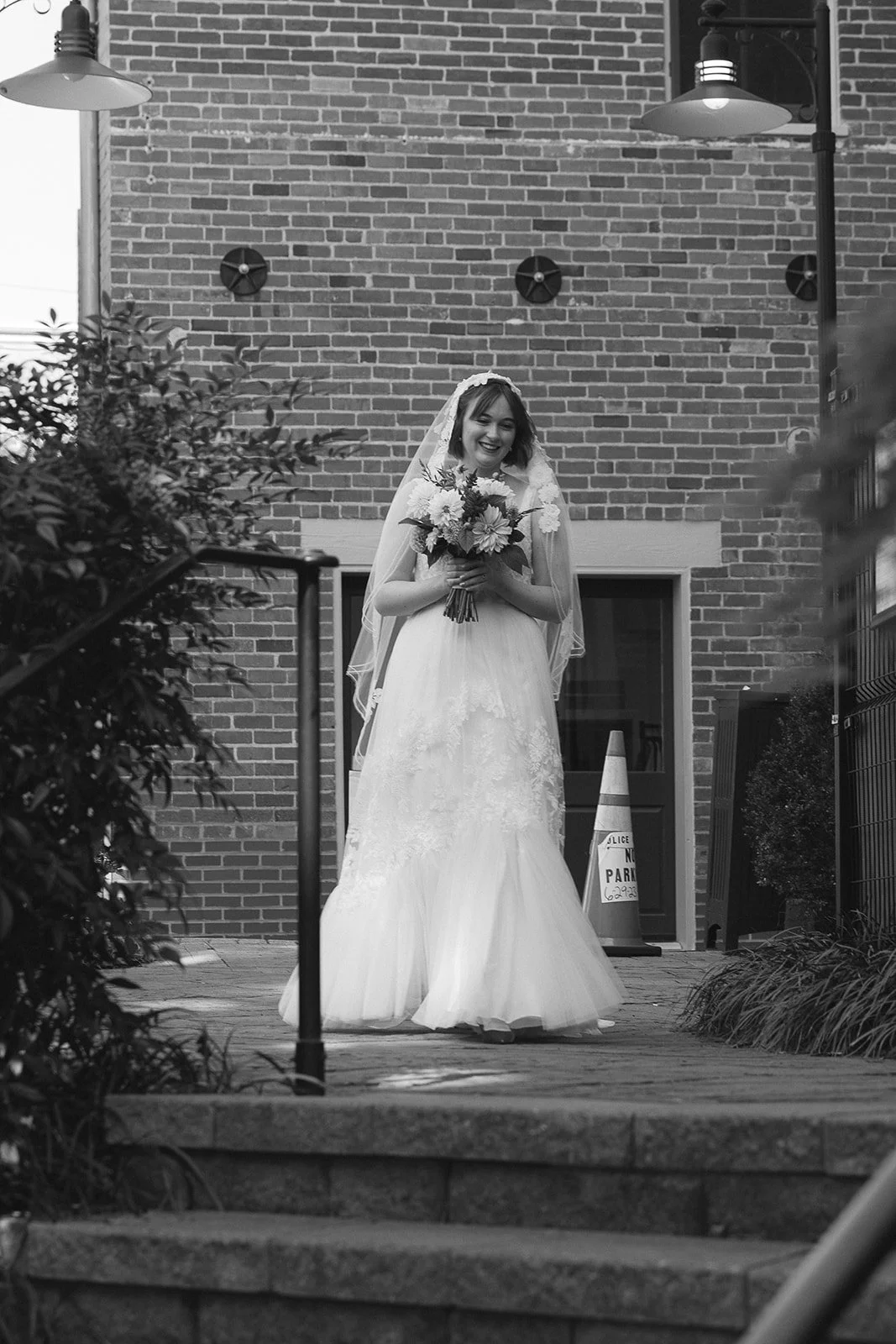 Black and white photo of a smiling bride in a wedding dress, holding a bouquet of flowers, standing outdoors in front of a brick building. Staunton Virginia Wedding Photographer.