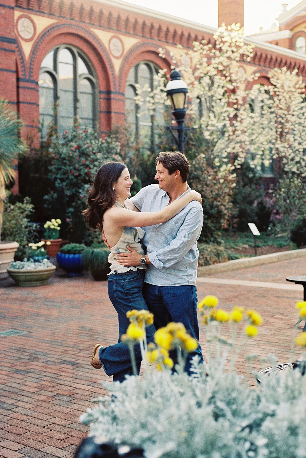 A couple dancing and smiling in an outdoor garden with blooming trees and flowering plants in the background.