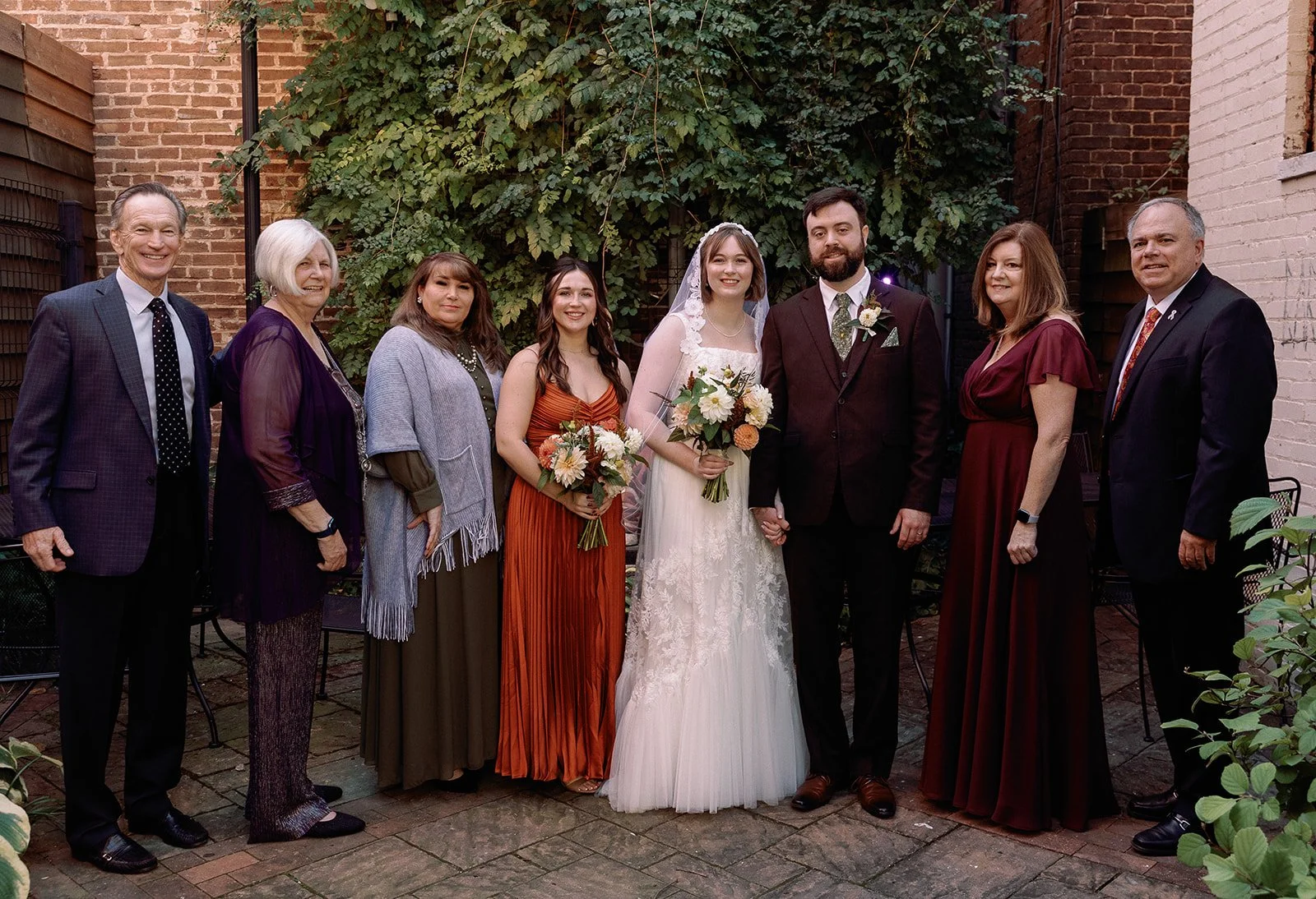 A wedding party of nine people standing outdoors against a brick wall with greenery. The bride and groom are in the center holding hands, with the bride wearing a white lace wedding gown and veil, and the groom in a dark suit. The others are dressed 