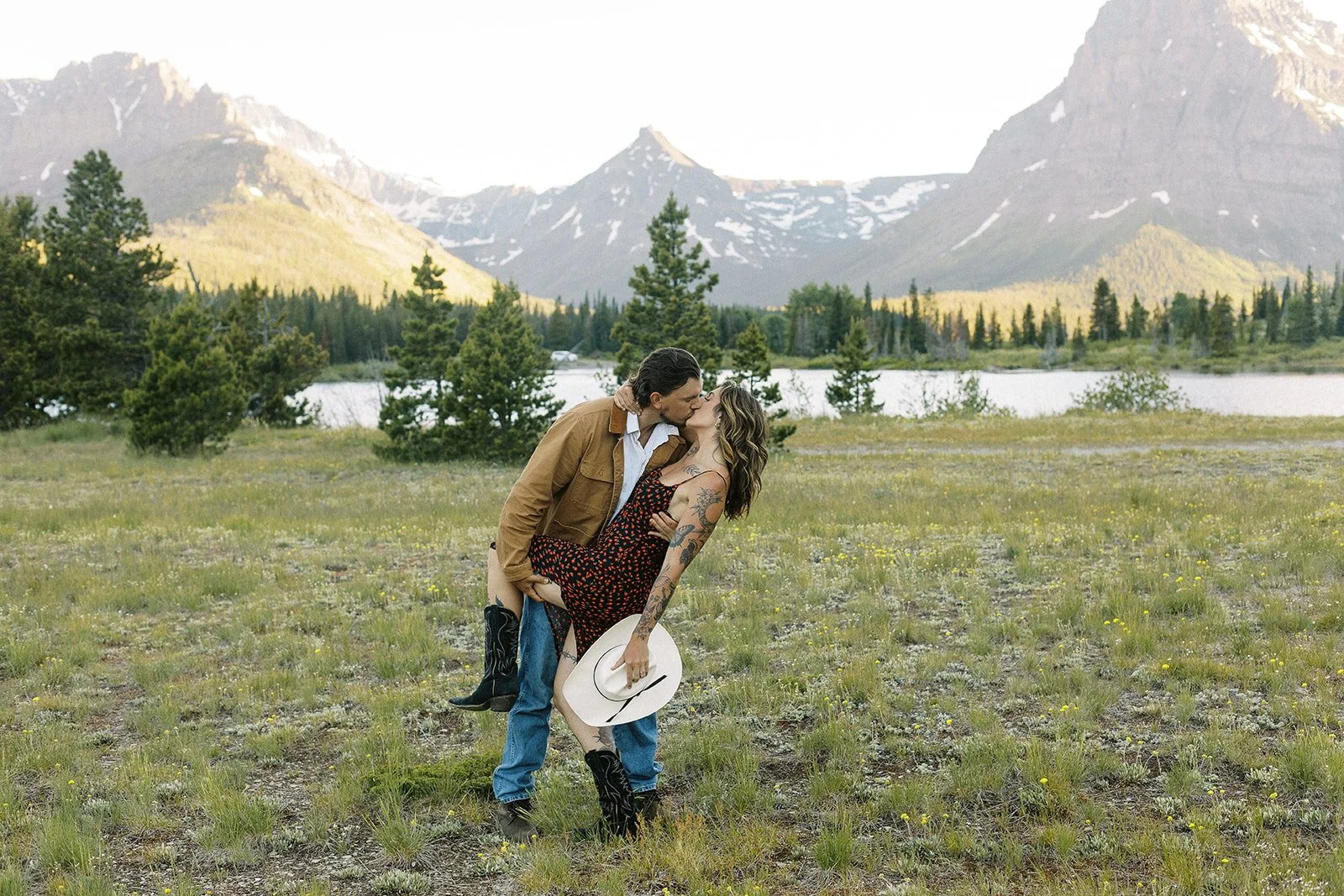 A couple sharing a kiss in a field with mountains, trees, and a body of water in the background.