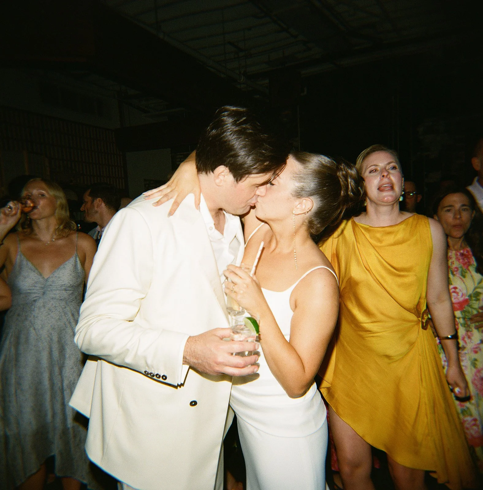 A man and woman are dancing closely at a party, with the man holding a drink and the woman holding a cigarette. They are surrounded by other partygoers, including a woman in a yellow dress and a woman in a gray dress eating a burger.