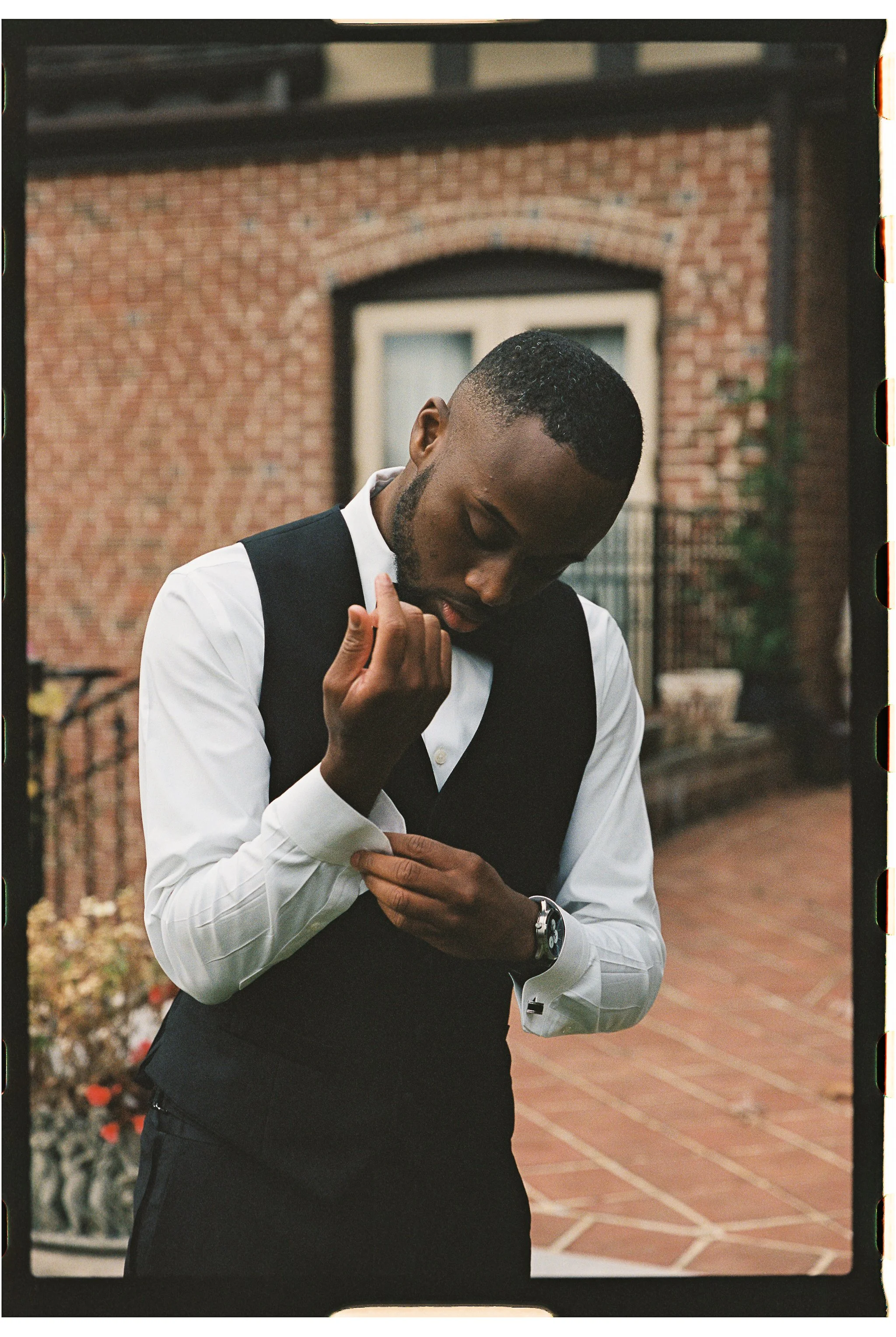 Man adjusting cufflink on white shirt and black vest, standing in front of brick building. gramercy mansion maryland wedding