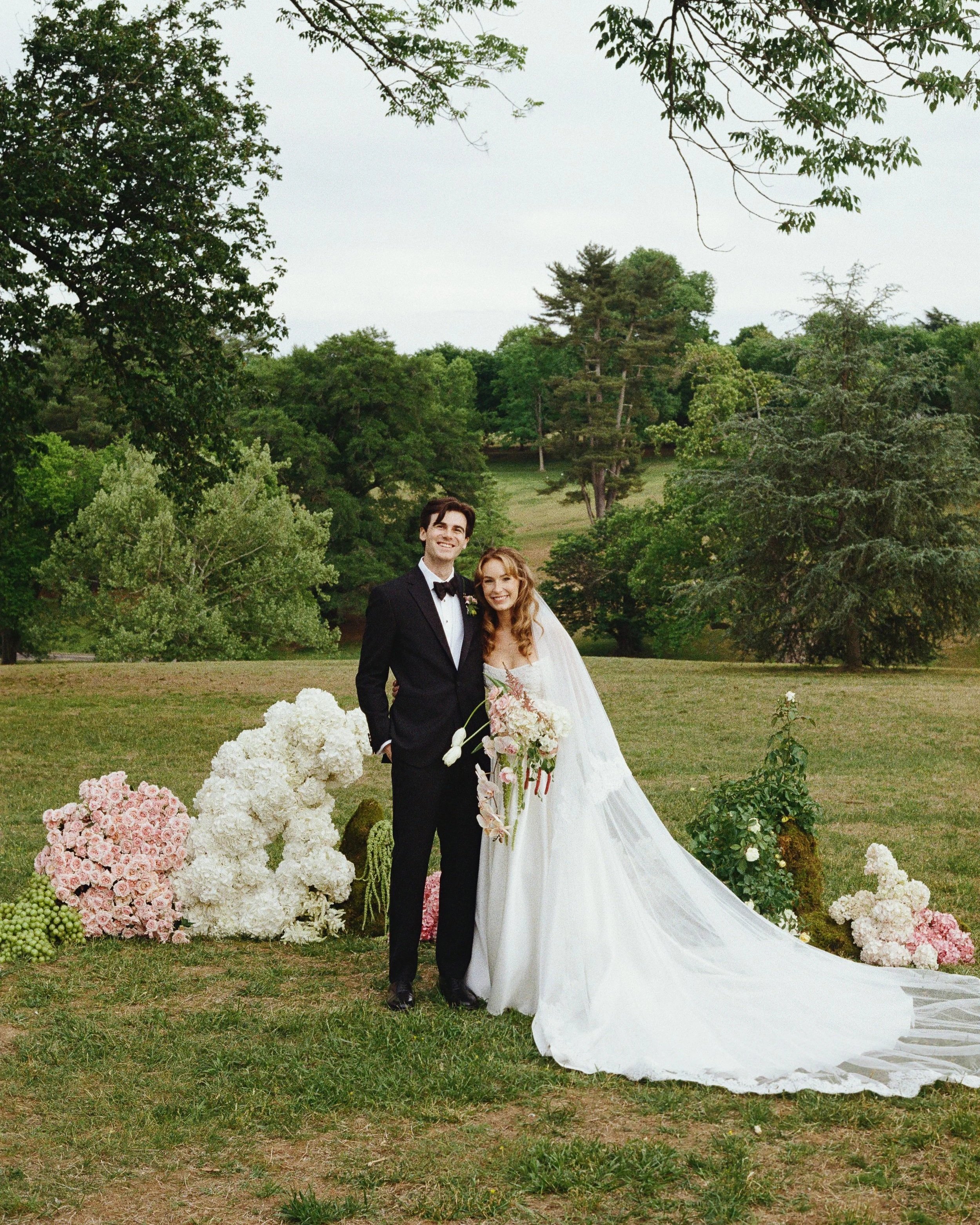 A newlywed couple standing outdoors in a park-like setting, with trees and grass in the background. The groom is wearing a black tuxedo and the bride is wearing a white wedding gown, holding a bouquet of flowers. They are smiling and posing for a photo, surrounded by white and pink floral arrangements.