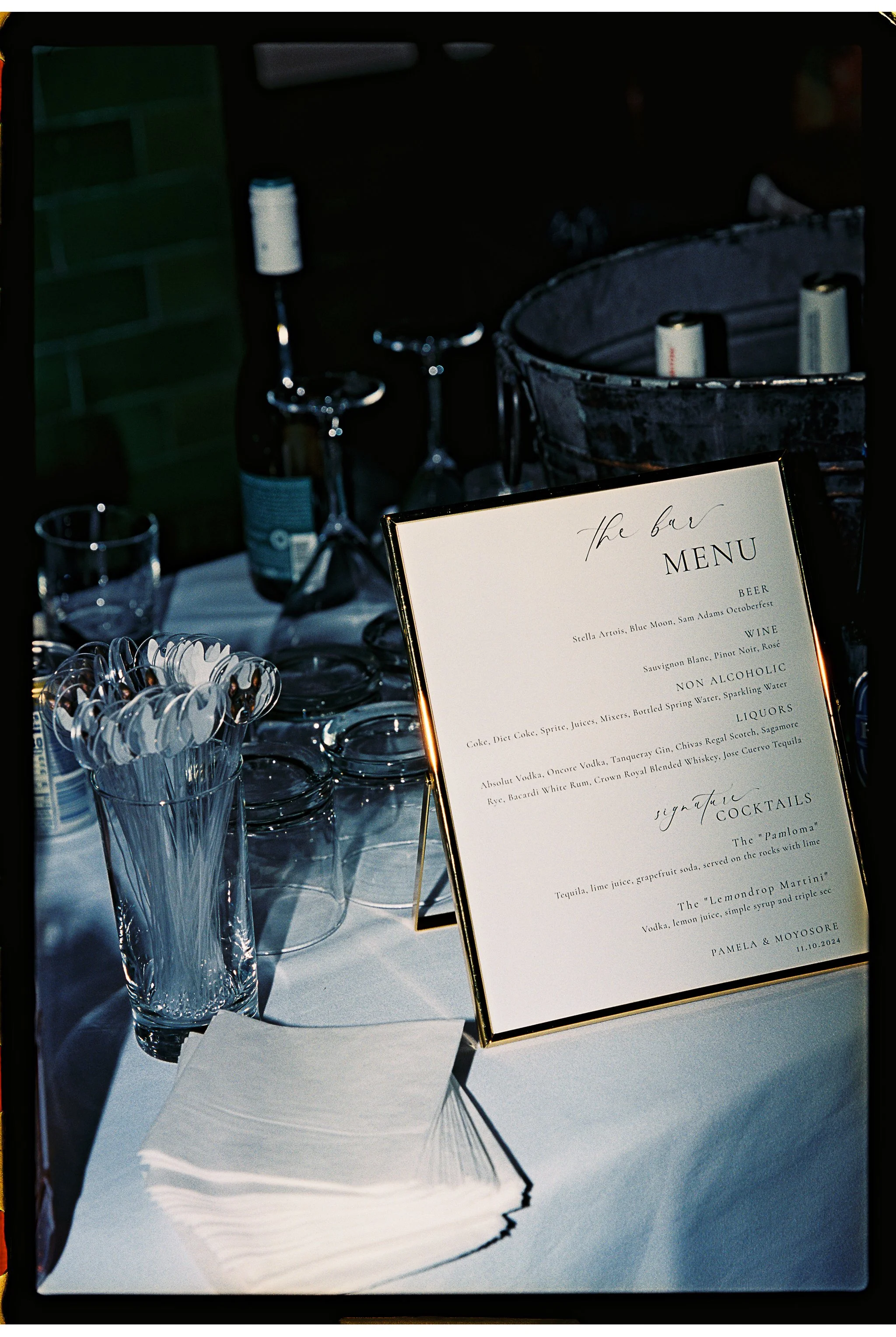 Close-up of a bar setup with a menu on a stand listing drinks, including beer, wine, non-alcoholic options, liquors, and signature cocktails. Surrounding items include stacked napkins, glasses, and a cup of stirrers. A metal bucket with bottles is al